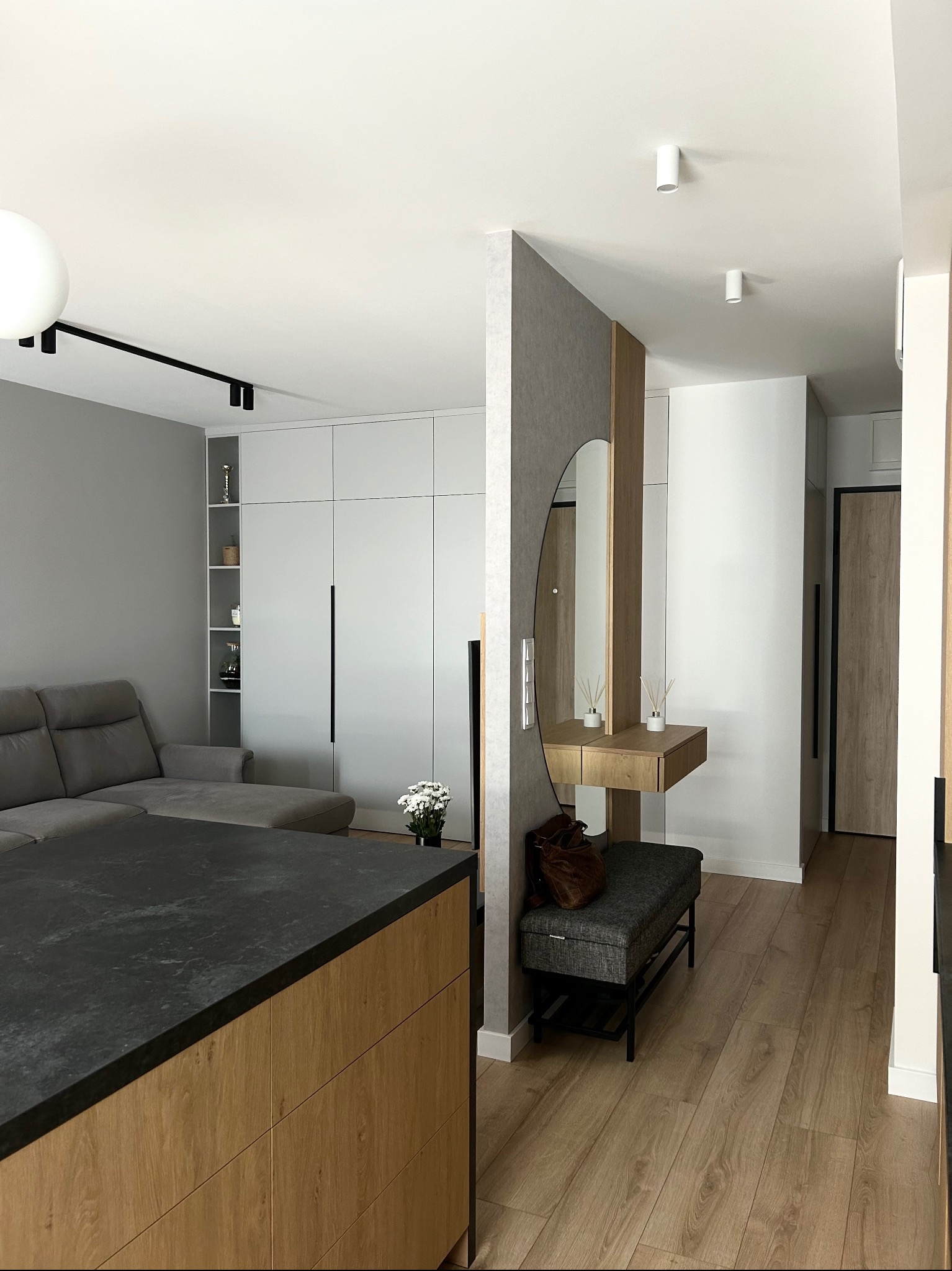 Wnętrze mieszkania z szarą sofą, białą szafą z czarnymi uchwytami, wyspą kuchenną z blatem z ciemnego kamienia, toaletką z okrągłym lustrem i drewnianym wykończeniem oraz siedziskiem obok