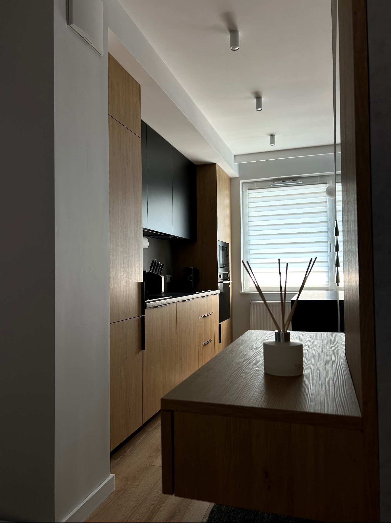 Nowoczesna kuchnia z drewnianymi frontami szafek dolnych i czarnymi górnymi, widok z perspektywy stołu z dyfuzorem zapachowym, jasne wnętrze z oknem.