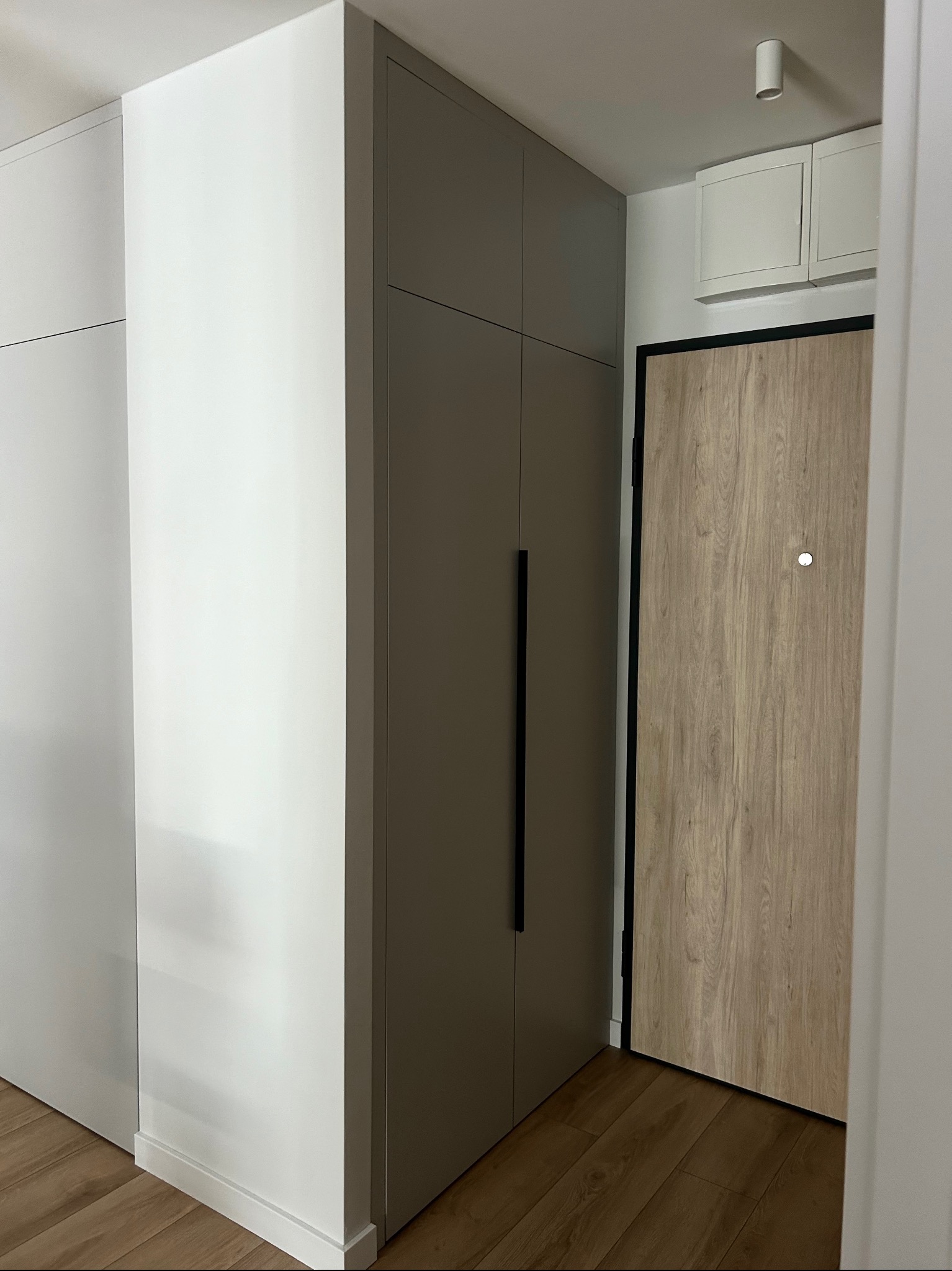 Wysoka, szara szafa z długimi, czarnymi uchwytami, wkomponowana we wnękę obok białych szafek, z drewnianą podłogą i drzwiami wejściowymi w tle.