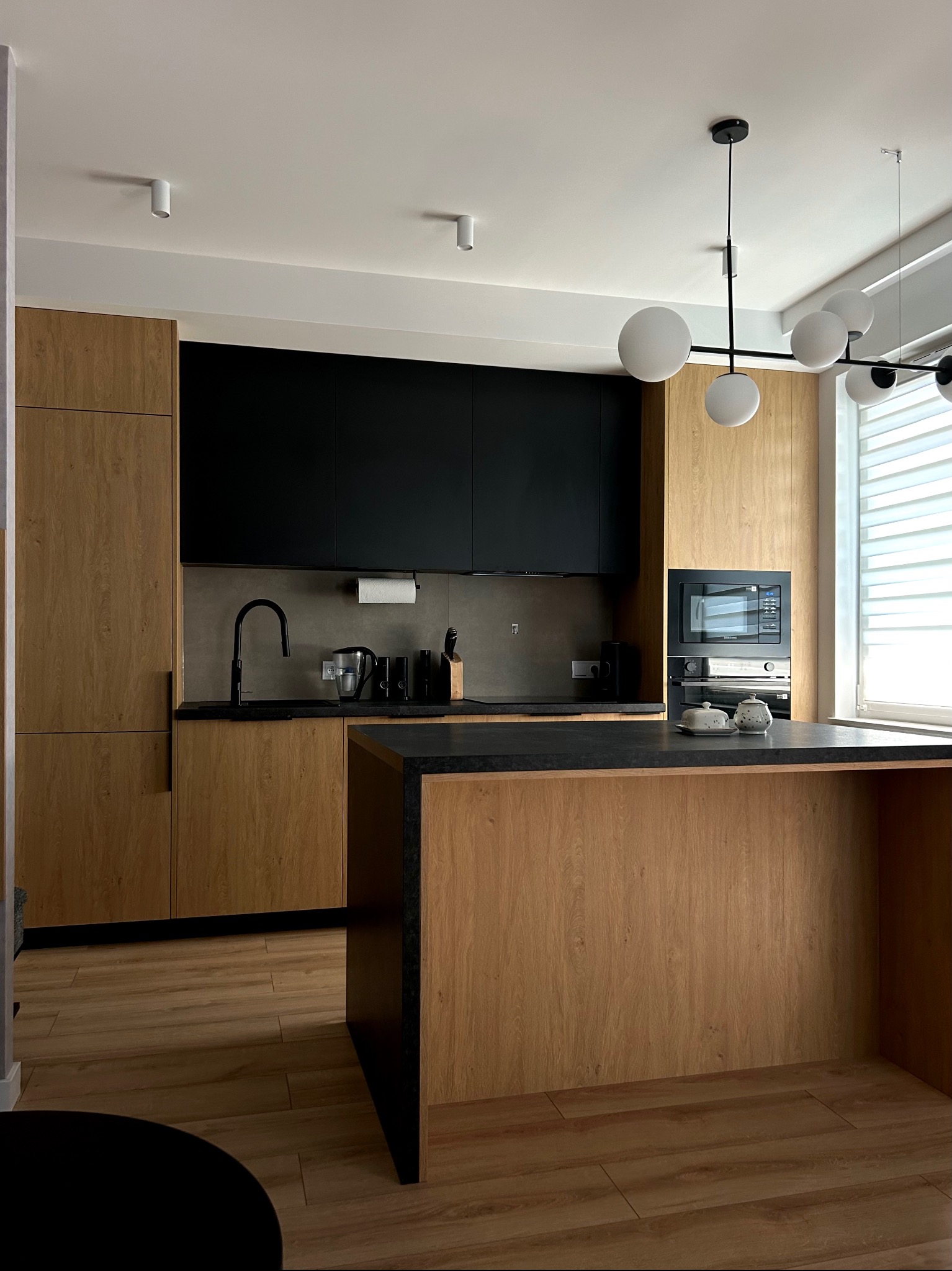 Nowoczesna kuchnia z drewnianymi frontami szafek, czarnymi elementami i wyspą kuchenną z blatem z czarnego kamienia, oświetlona lampą wiszącą z kulistymi kloszami.