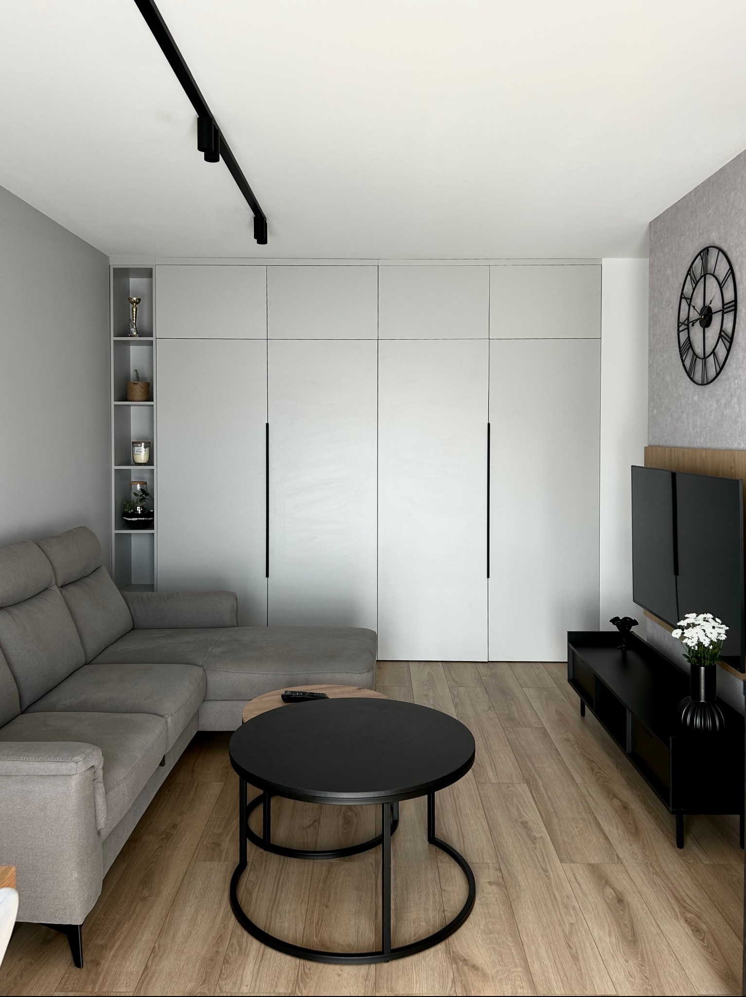 Nowoczesny salon z szarą sofą narożną, okrągłym czarnym stolikiem kawowym i dużą białą szafą wnękową z minimalistycznymi uchwytami, uzupełniony o czarny zegar na ścianie i oświetlenie szynowe