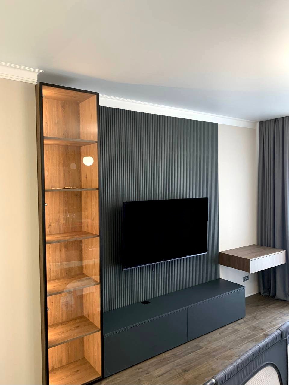 Nowoczesny salon z zabudową meblową w odcieniach szarości i drewna, obejmującą wysoką przeszkloną witrynę z oświetleniem, szafkę RTV pod telewizorem na tle ściany z pionowymi panelami oraz wiszący...