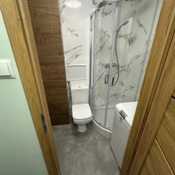 MAIBUD ADAM MROZEK - Wykończona łazienka z kabiną prysznicową, toaletą i pralką, widok zza otwartych drewnianych drzwi. Płytki imitujące marmur na ścianach, szare płytki na podłodze.