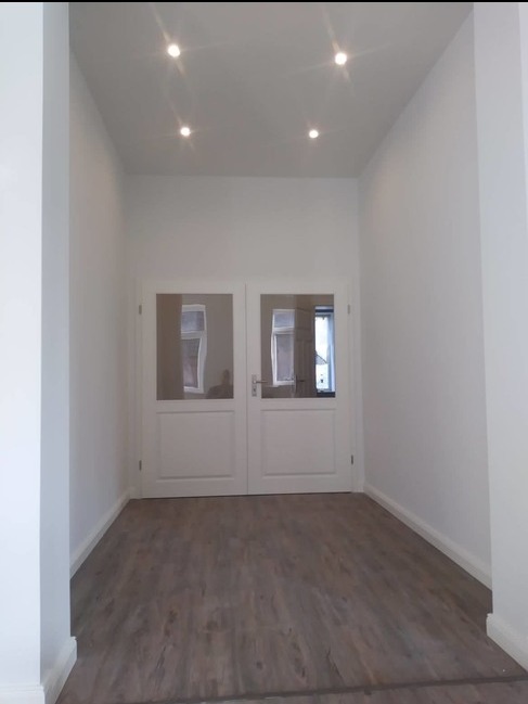 Korytarz po remoncie w Łodzi: świeżo pomalowane białe ściany, nowe panele podłogowe imitujące drewno i białe dwuskrzydłowe drzwi z przeszkleniami.