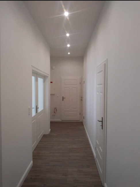 Korytarz po remoncie z białymi ścianami, drewnopodobną podłogą i oświetleniem punktowym w suficie.