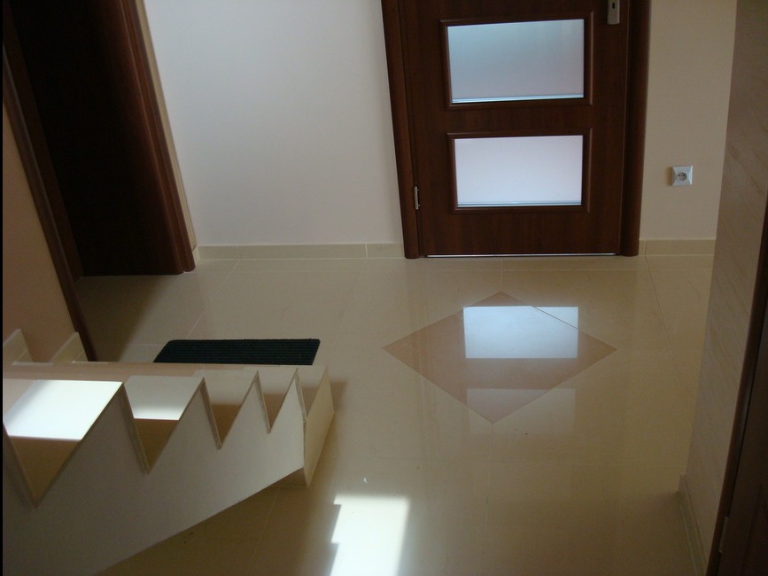 Beżowe płytki podłogowe z dekoracyjnym rombem, białe schody z płyty karton gips, ciemne drzwi z szybami i gniazdko elektryczne na ścianie.