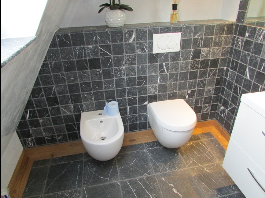 Wykończona łazienka z czarnymi marmurkowymi płytkami, białym bidetem z niebieską rolką papieru toaletowego, wiszącą toaletą i drewnianymi listwami przypodłogowymi.