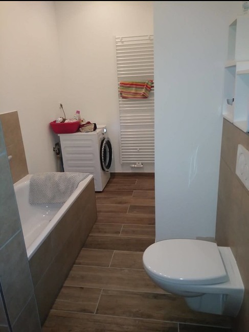 Nowoczesna łazienka z brązowymi płytkami imitującymi drewno, białą wanną z szarą ścierką, toaletą podwieszaną i pralką z czerwonym koszem na pranie. Grzejnik drabinkowy z ręcznikiem w tle.