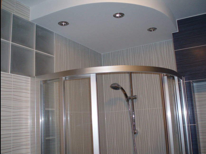 Zaokrąglona kabina prysznicowa z chromowaną armaturą i deszczownicą, białe płytki ścienne z delikatnym wzorem fal, podwieszany sufit z oświetleniem punktowym.