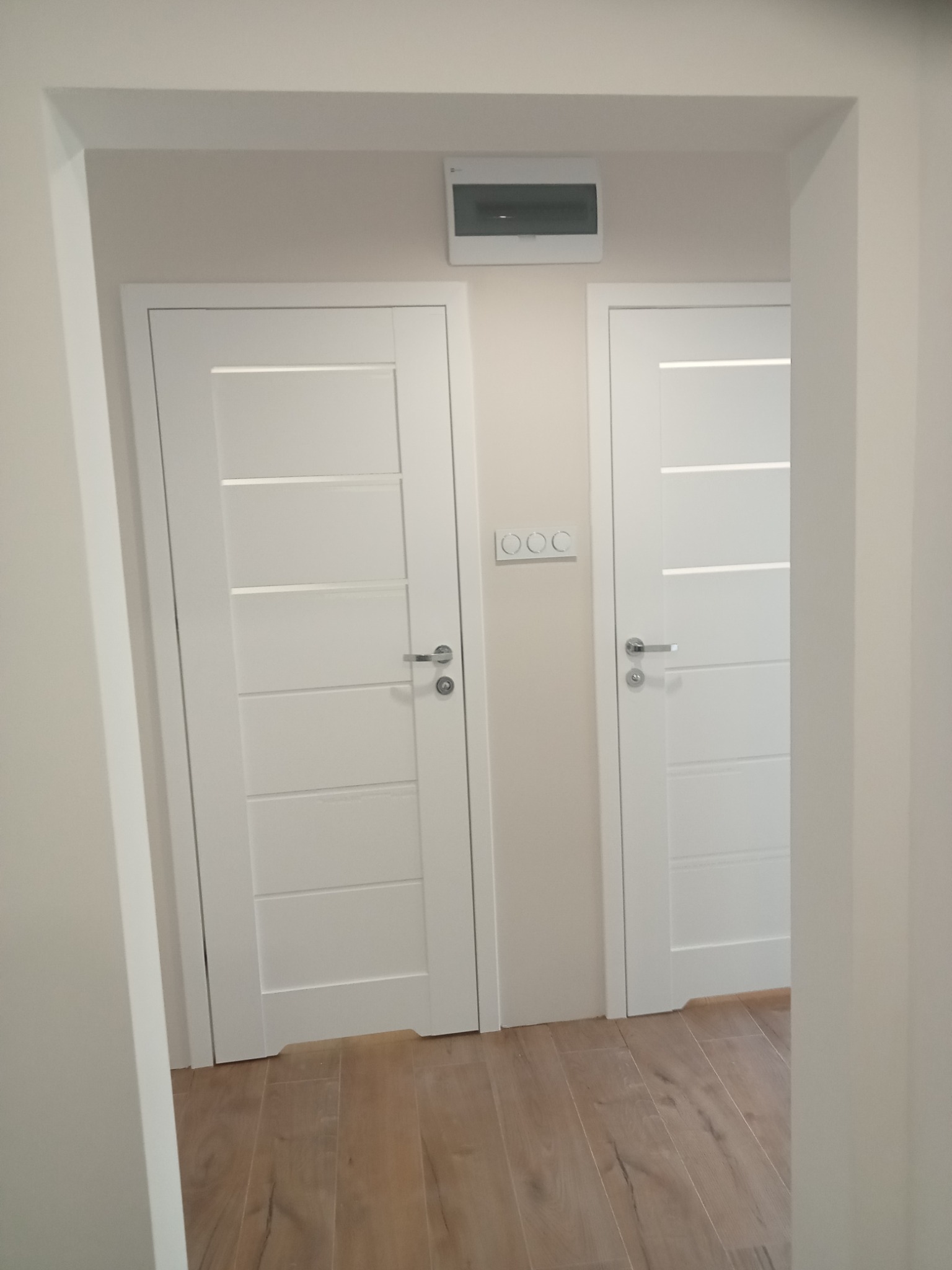 Dwa białe, nowoczesne drzwi z poziomymi przeszkleniami, zamontowane w jasnym korytarzu z panelami podłogowymi. Widoczne włączniki światła.