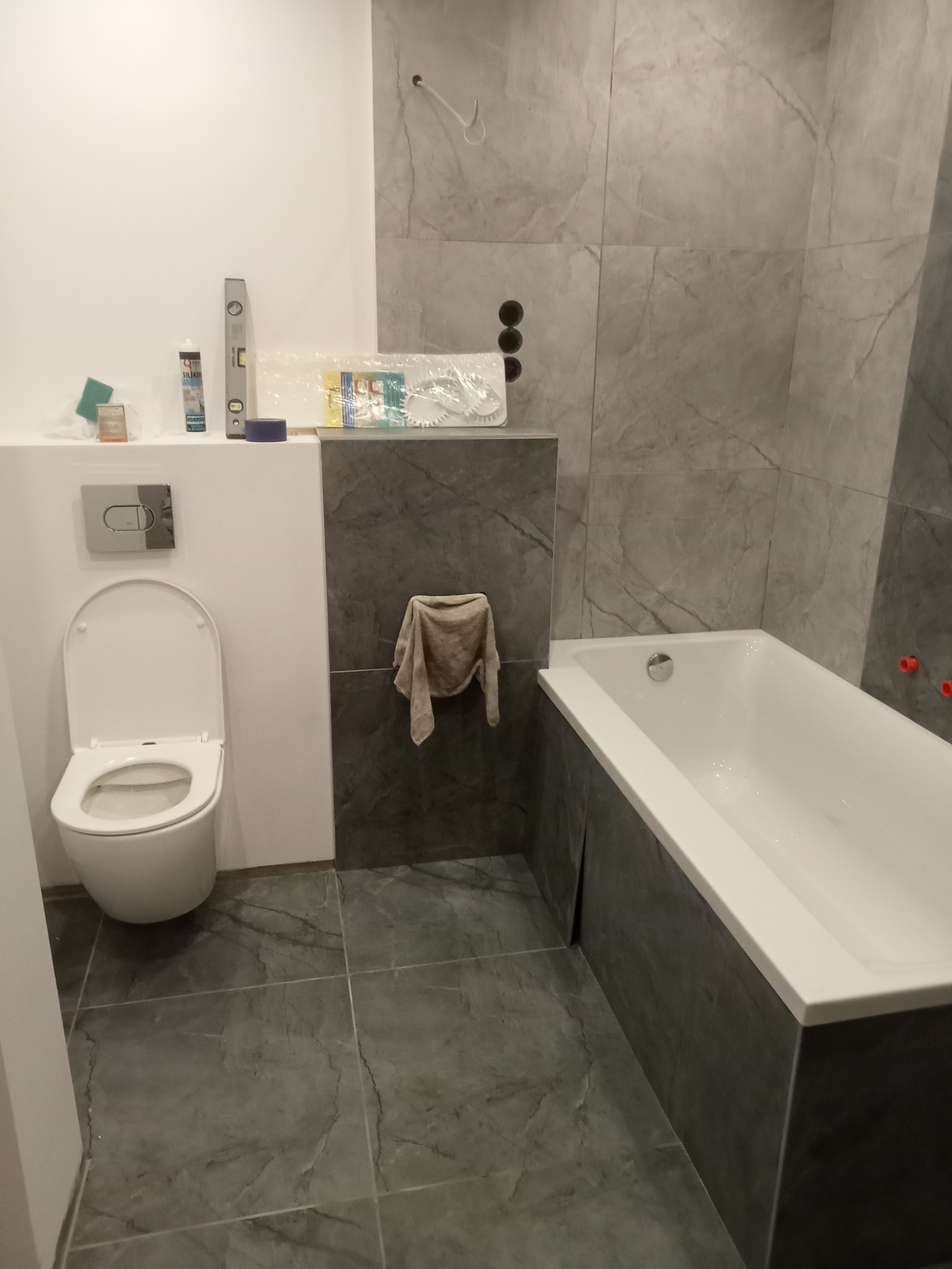 Szara łazienka w trakcie wykańczania: widoczna wanna, toaleta, płytki imitujące marmur. Na półce akcesoria i poziomica. Na ścianie przyłącza. Ręcznik zawieszony na ścianie.