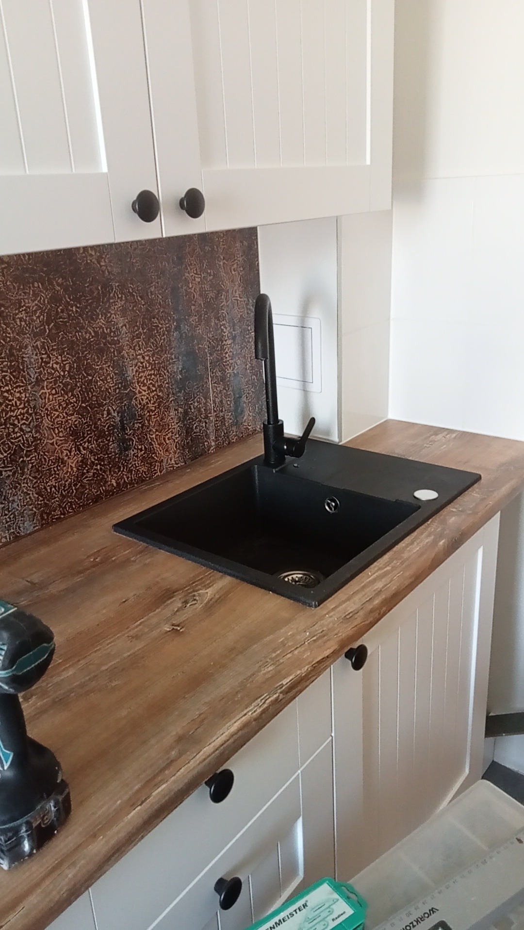 Nowoczesna kuchnia z czarnym zlewem i baterią, drewnianym blatem oraz białymi szafkami. Widoczna wiertarka akumulatorowa.