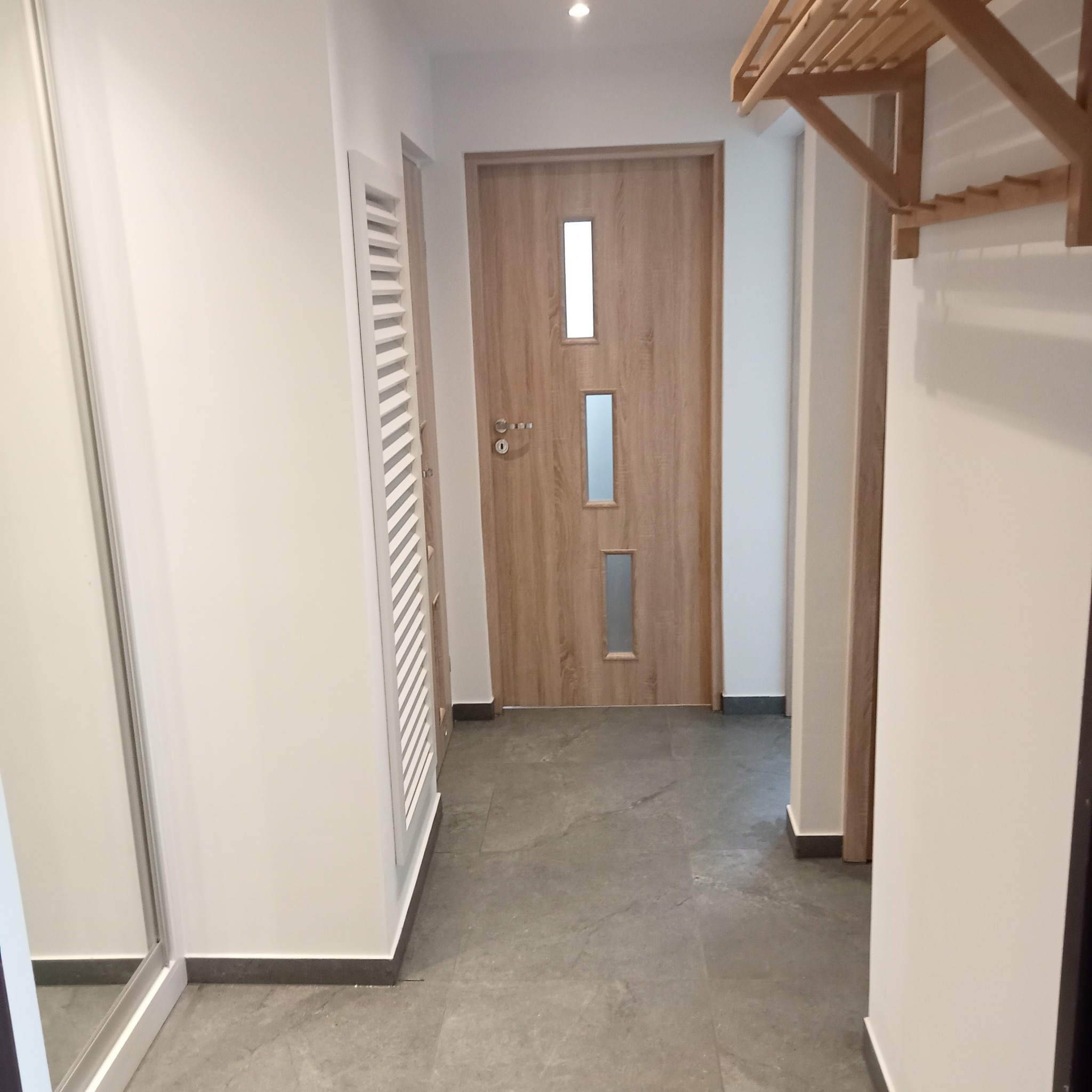 Jasne wnętrze przedpokoju z drewnianą okleiną drzwi, szarą podłogą i białymi ścianami. Widoczny grzejnik dekoracyjny i wieszak na ubrania.