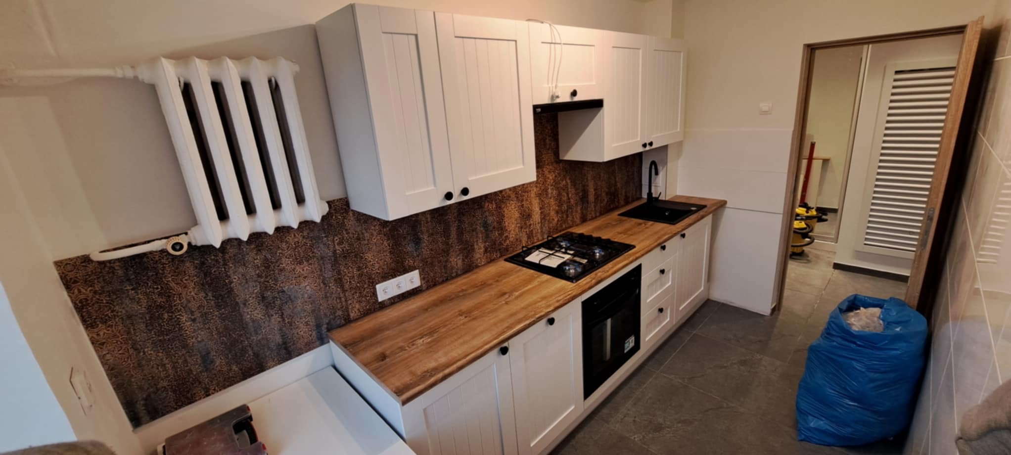 Wykończona kuchnia z białą zabudową i drewnianym blatem. Na ścianie dekoracyjna tapeta. Widoczny grzejnik żeberkowy i sprzęt AGD w zabudowie.