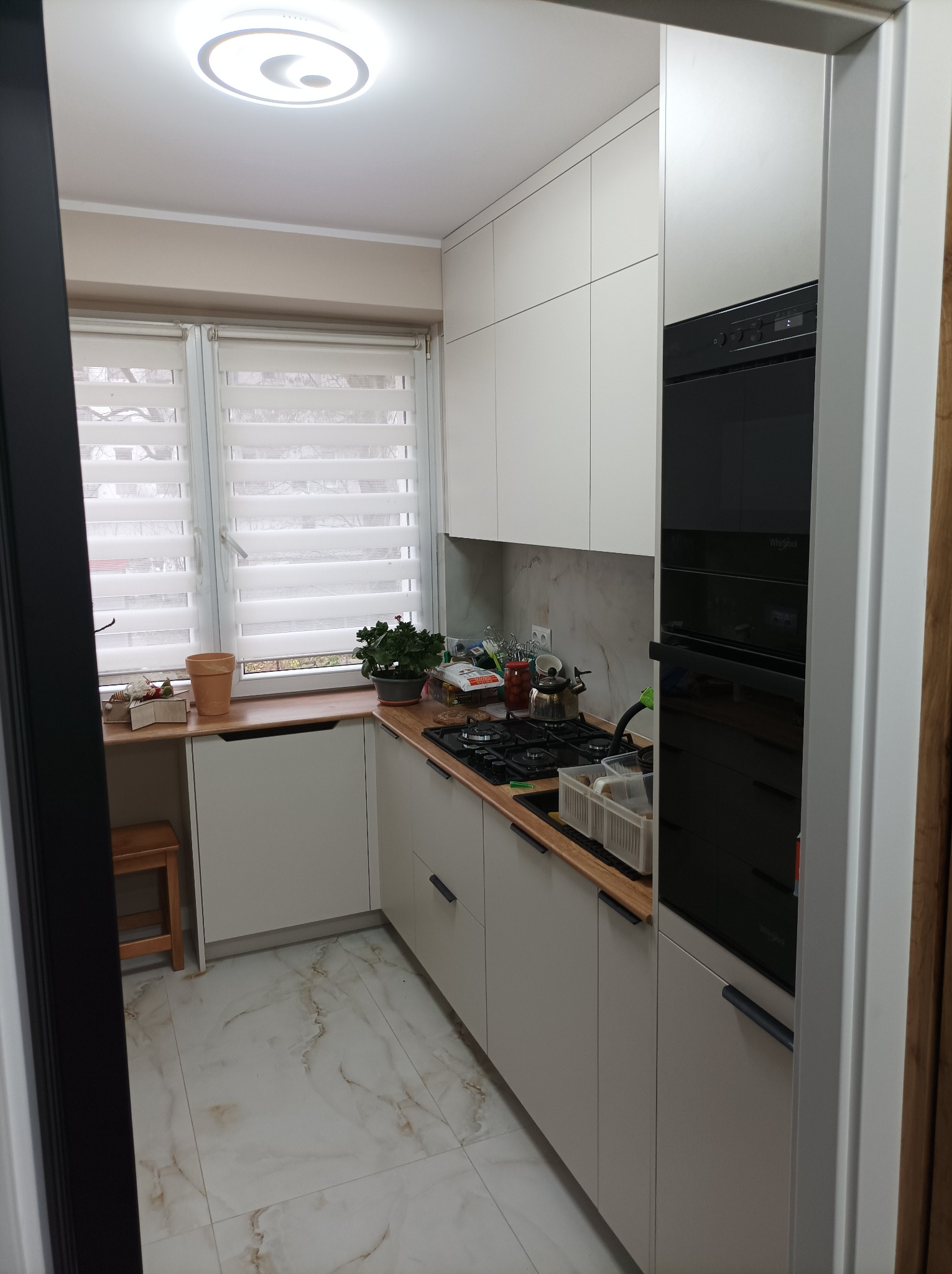 Jasna kuchnia z drewnianym blatem i marmurową podłogą, widziana zza framugi drzwi. Okno z żaluzjami w tle, nowoczesne szafki i wbudowany piekarnik.