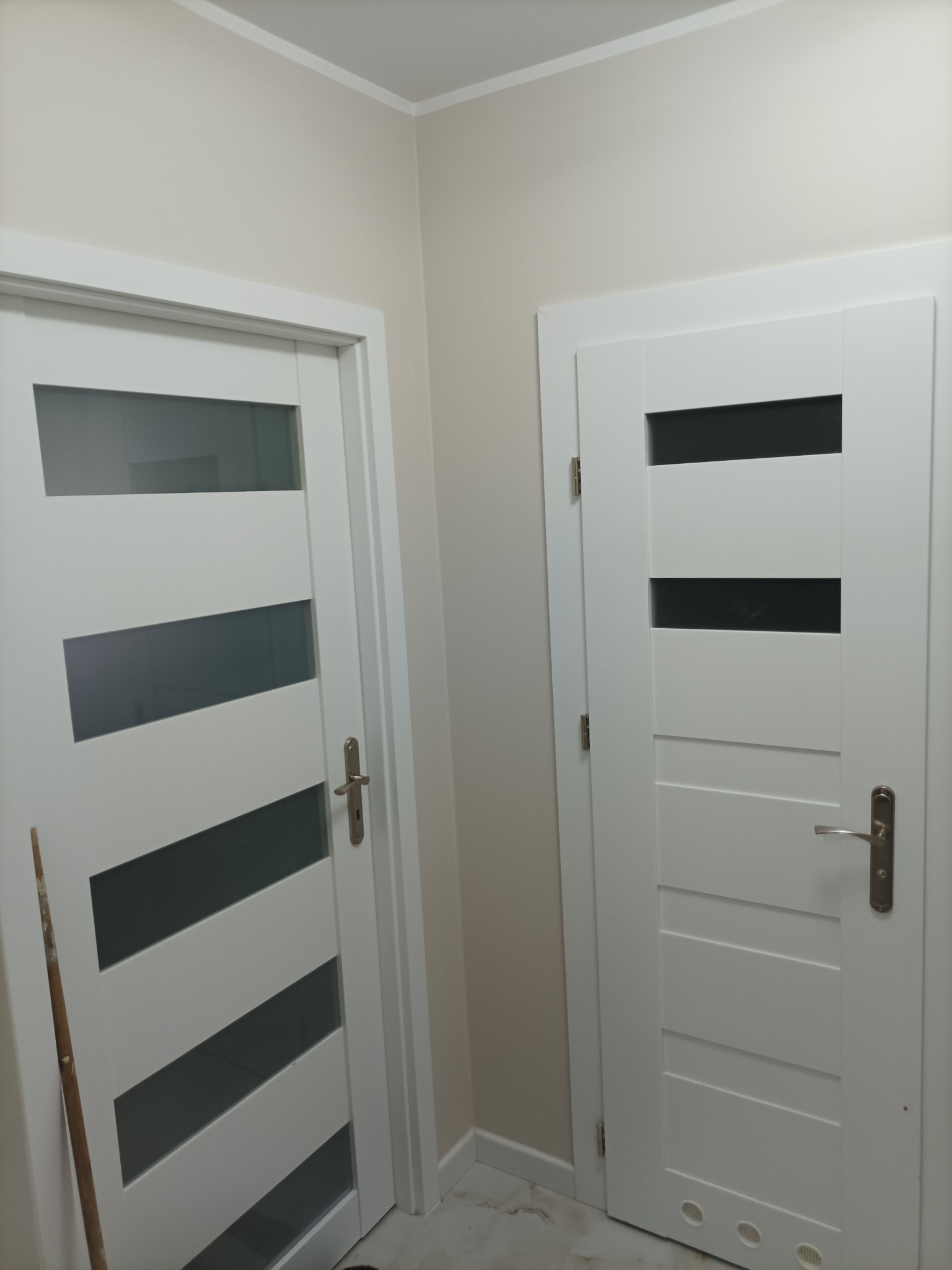 Dwa białe, nowoczesne drzwi wewnętrzne z przeszkleniami, zamontowane w jasnym pomieszczeniu. Po lewej drzwi z trzema poziomymi szybami, po prawej z czterema.