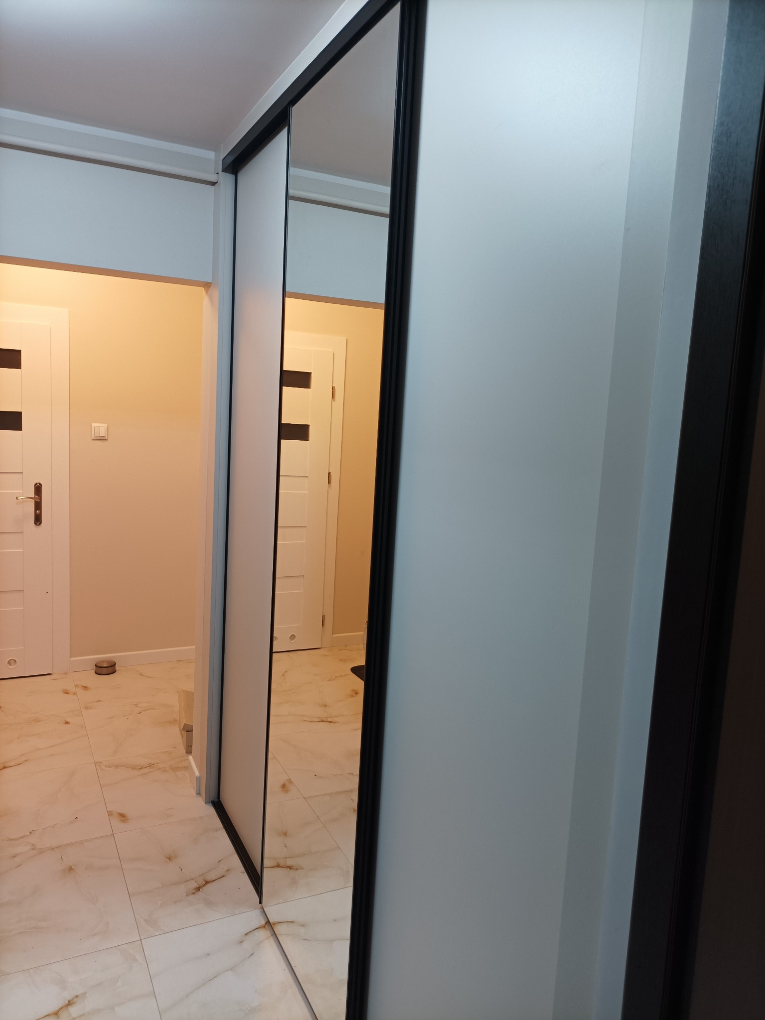 Nowoczesna szafa przesuwna z lustrem w przedpokoju, odbijająca białe drzwi i marmurowe płytki podłogowe. Minimalistyczny design z czarnymi obramowaniami.