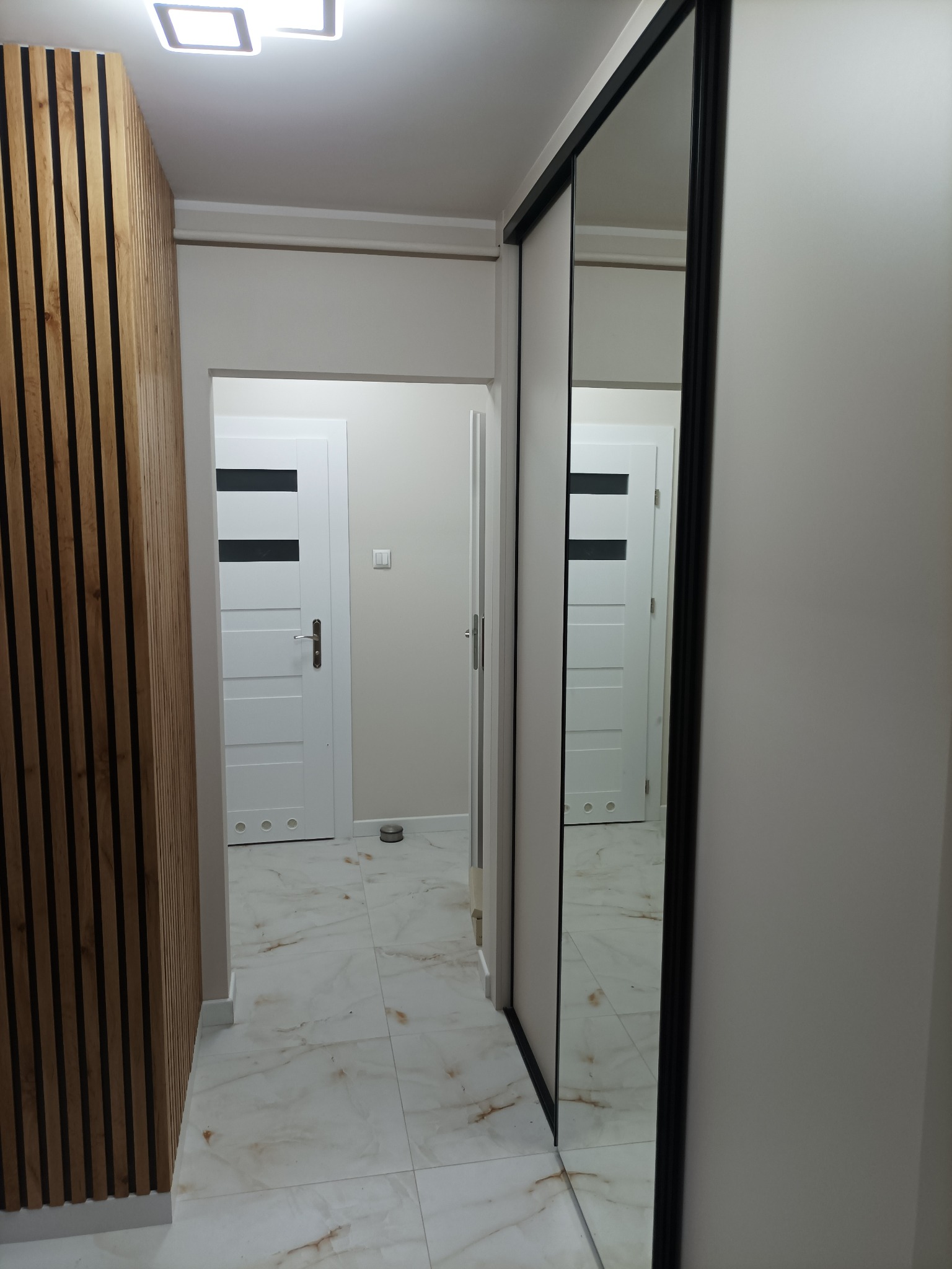 Nowoczesny korytarz z drewnianą ścianą, lustrzaną szafą i marmurową podłogą. Widoczne białe drzwi z czarnymi akcentami i minimalistyczne oświetlenie sufitowe.