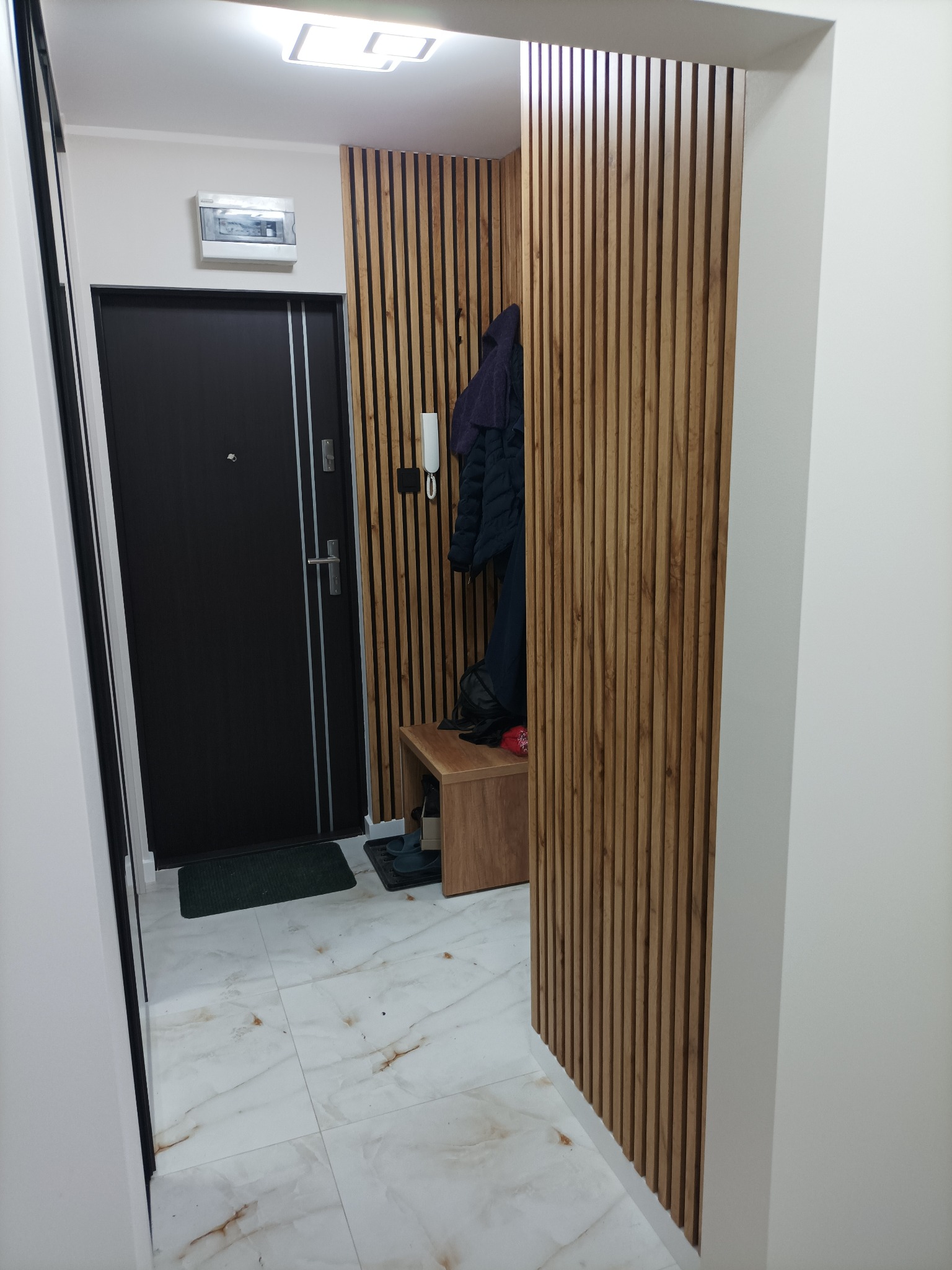 Nowoczesny przedpokój z czarnymi drzwiami, panelami ściennymi z drewna i marmurową podłogą. Widoczna szafka na buty i wieszak na ubrania. Oświetlenie sufitowe.