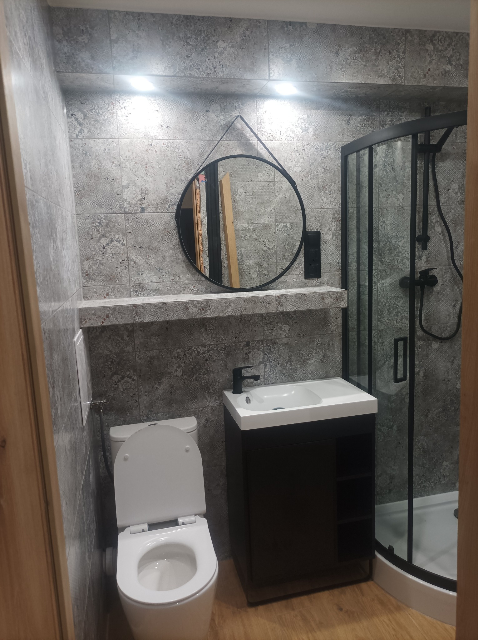 Wykończona łazienka z kabiną prysznicową, umywalką z szafką i toaletą. Ściany wyłożone szarymi płytkami z wzorem. Okrągłe lustro i czarne akcenty.