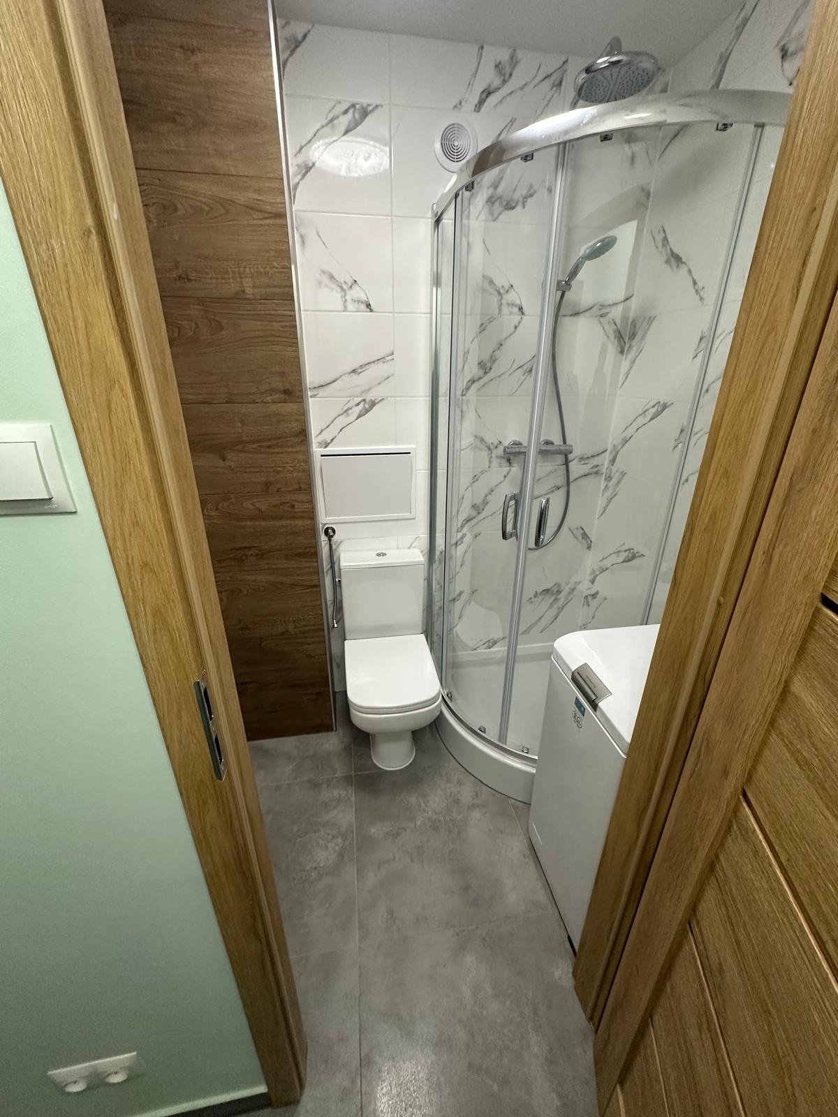 Wykończona łazienka z kabiną prysznicową, toaletą i pralką, widok zza otwartych drewnianych drzwi. Płytki imitujące marmur na ścianach, szare płytki na podłodze.
