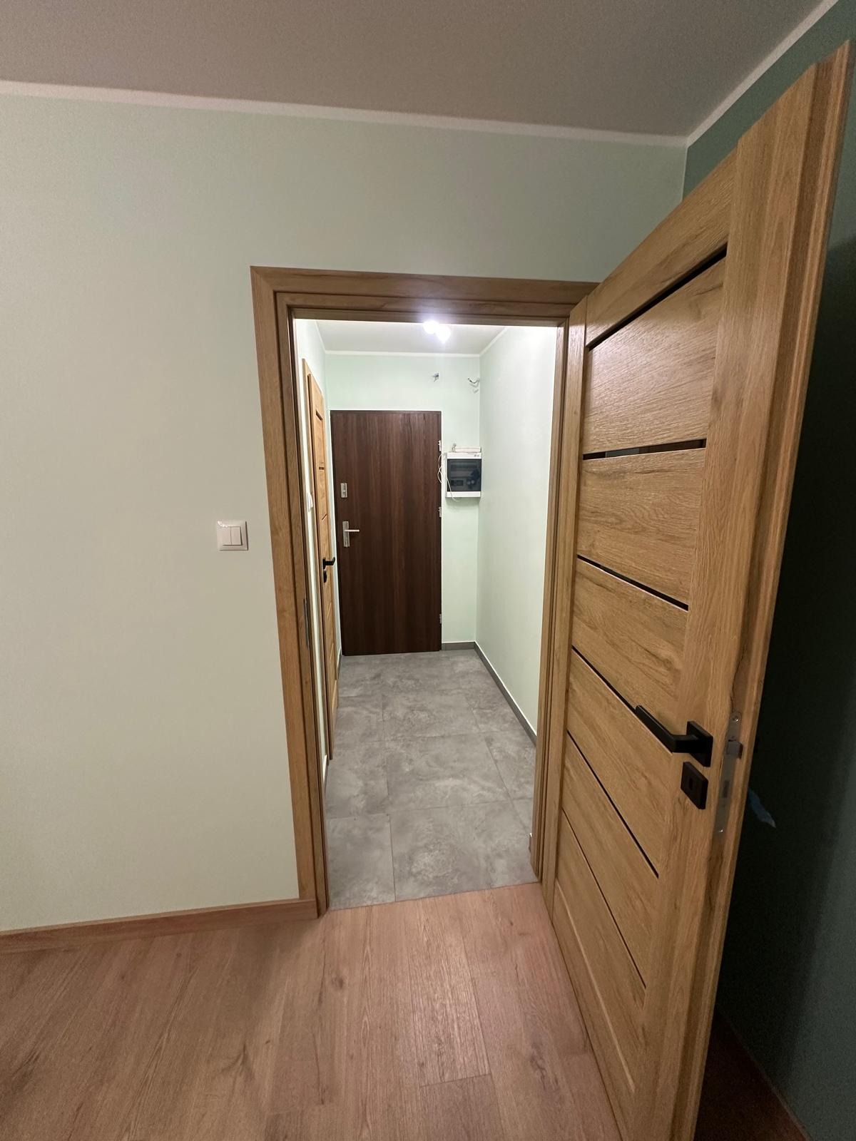 Otwarta, drewniana, nowoczesna dębowa okleina drzwi wewnętrznych z czarną klamką, widok na korytarz z szarą podłogą i brązowymi drzwiami wejściowymi.