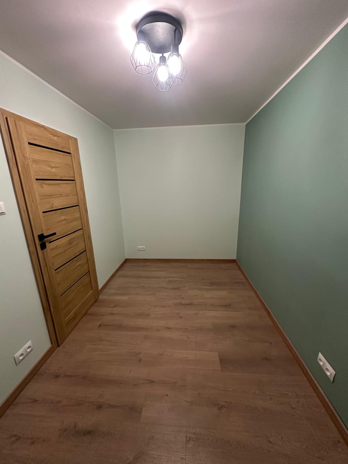 Pokój po remoncie w Dmosinie: drewniana podłoga, pomalowane ściany na zielono i jasnozielono, nowoczesna lampa sufitowa, drewniane drzwi z czarnymi akcentami.