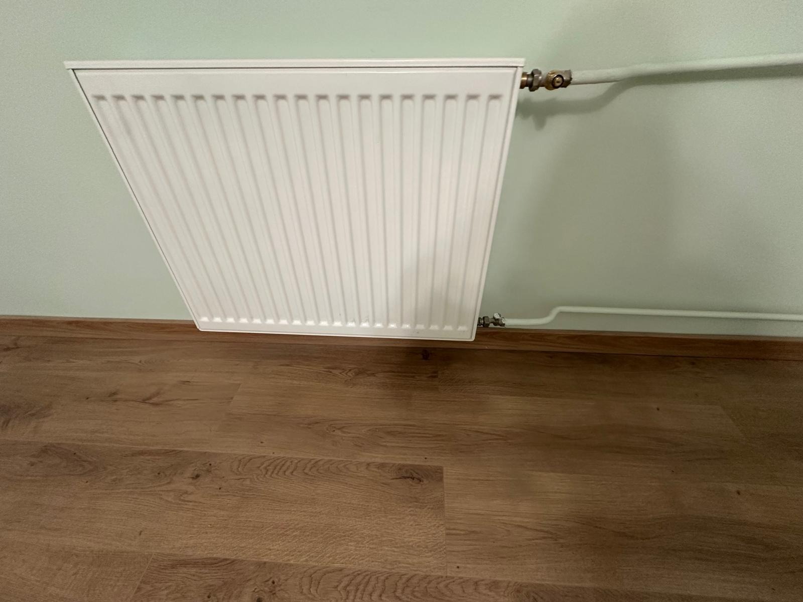 Biały, nowy grzejnik panelowy zamontowany na ścianie w pokoju z drewnianą podłogą. Widoczne rury doprowadzające wodę. Ściana pomalowana na jasnozielony kolor.