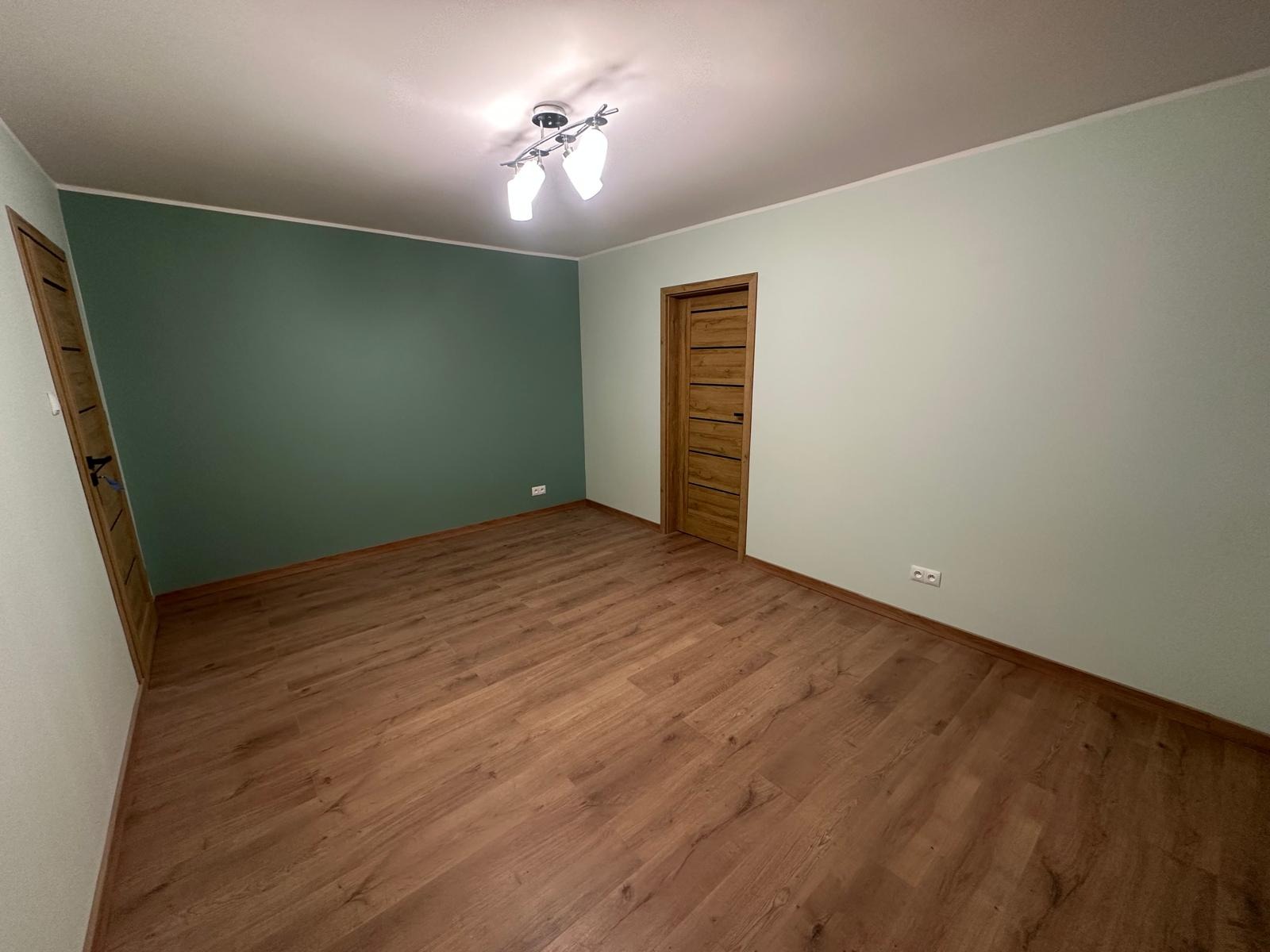 Pokój po remoncie: brązowe panele, ściany w odcieniach zieleni, drewniane drzwi z poziomymi żłobieniami, nowoczesna lampa sufitowa. Widok na odnowione wnętrze.