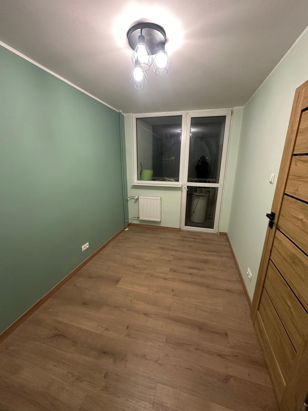 Jasne panele podłogowe w pokoju z zielonymi ścianami, nowoczesna lampa sufitowa i drewniane drzwi. Widok na balkon z grzejnikiem.