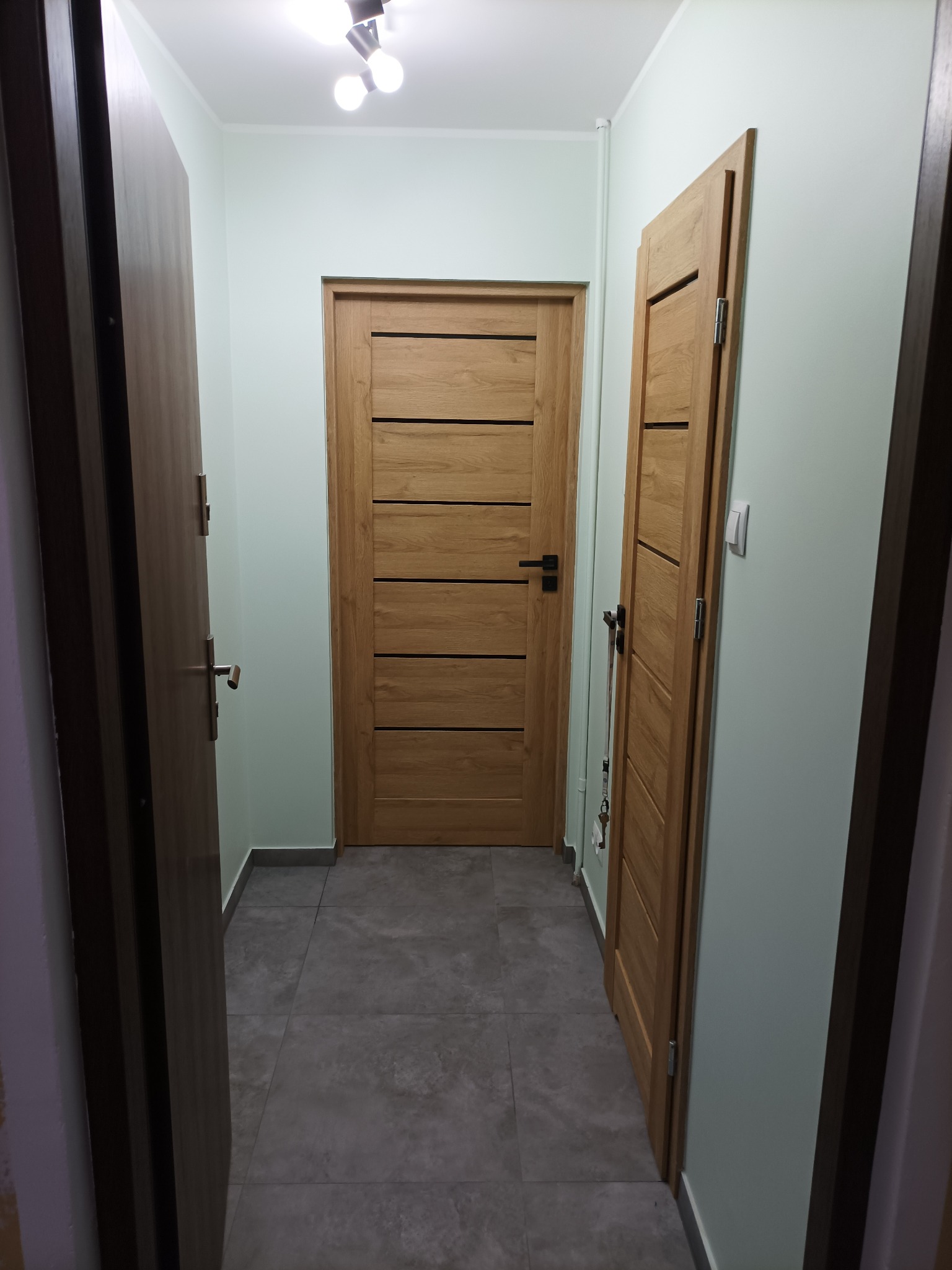 Korytarz z trzema drewnianymi drzwiami o nowoczesnym designie, szare płytki podłogowe, oświetlenie sufitowe. Widok z perspektywy wejścia.