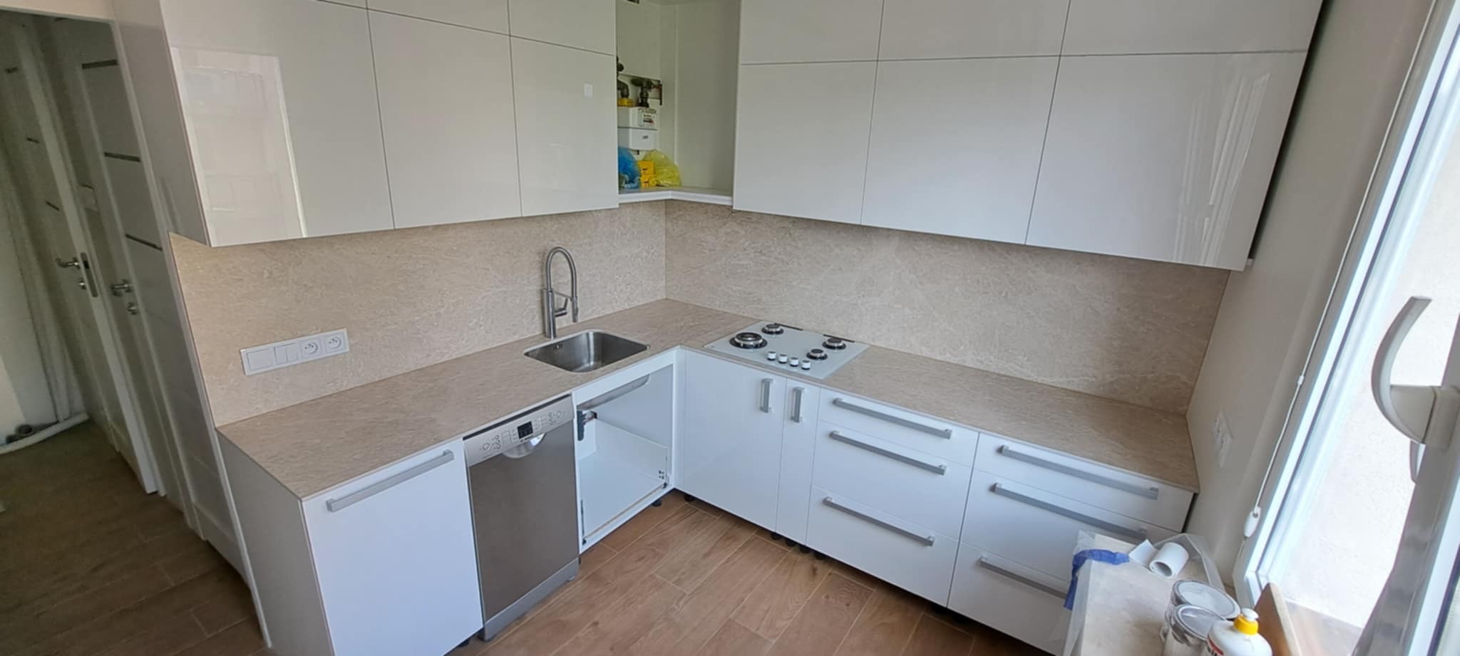 Nowoczesna, jasna kuchnia z białymi szafkami i beżowym blatem. Widoczny zlew, płyta grzewcza i drewniana podłoga. Minimalistyczny design z elementami wykończenia wnętrz.