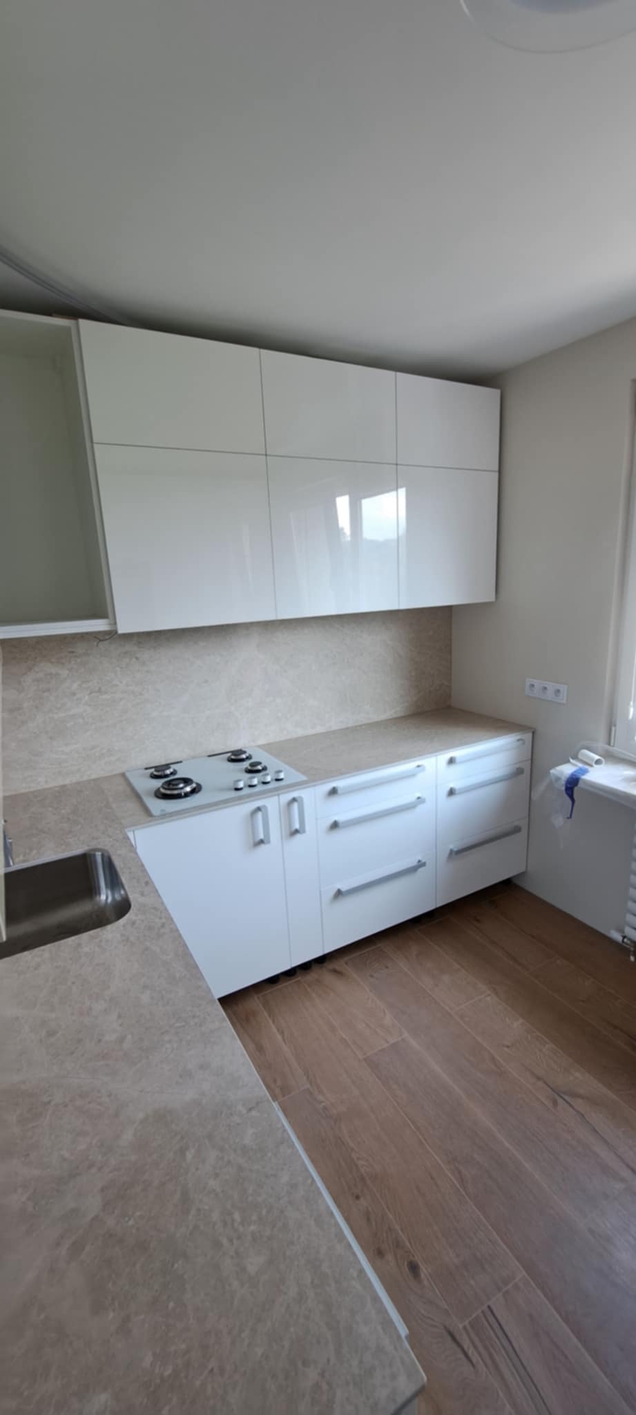 Nowoczesna kuchnia z białymi, błyszczącymi szafkami i blatem imitującym marmur. Widoczna płyta grzewcza i drewnopodobna podłoga. Minimalistyczny design.