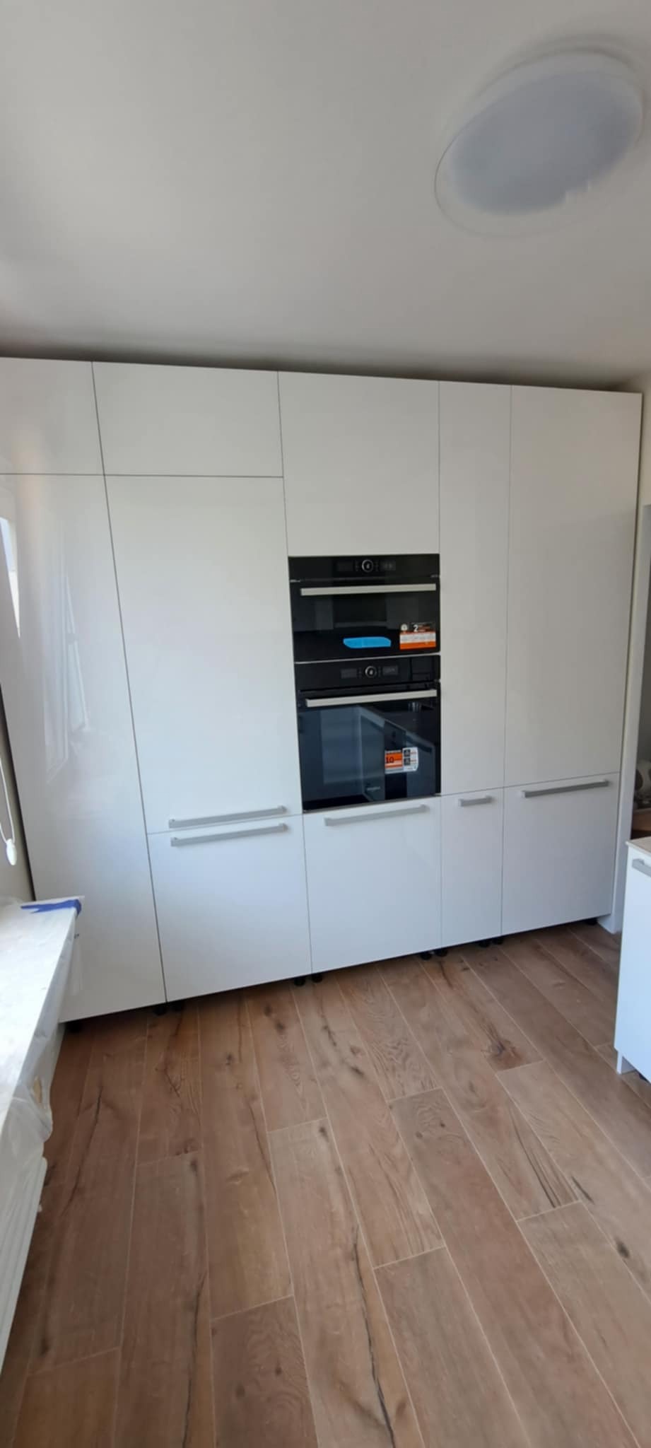 Biała zabudowa kuchenna z piekarnikiem i mikrofalówką, drewniana podłoga. Minimalistyczny design, nowoczesne wykończenie. Oświetlenie sufitowe.