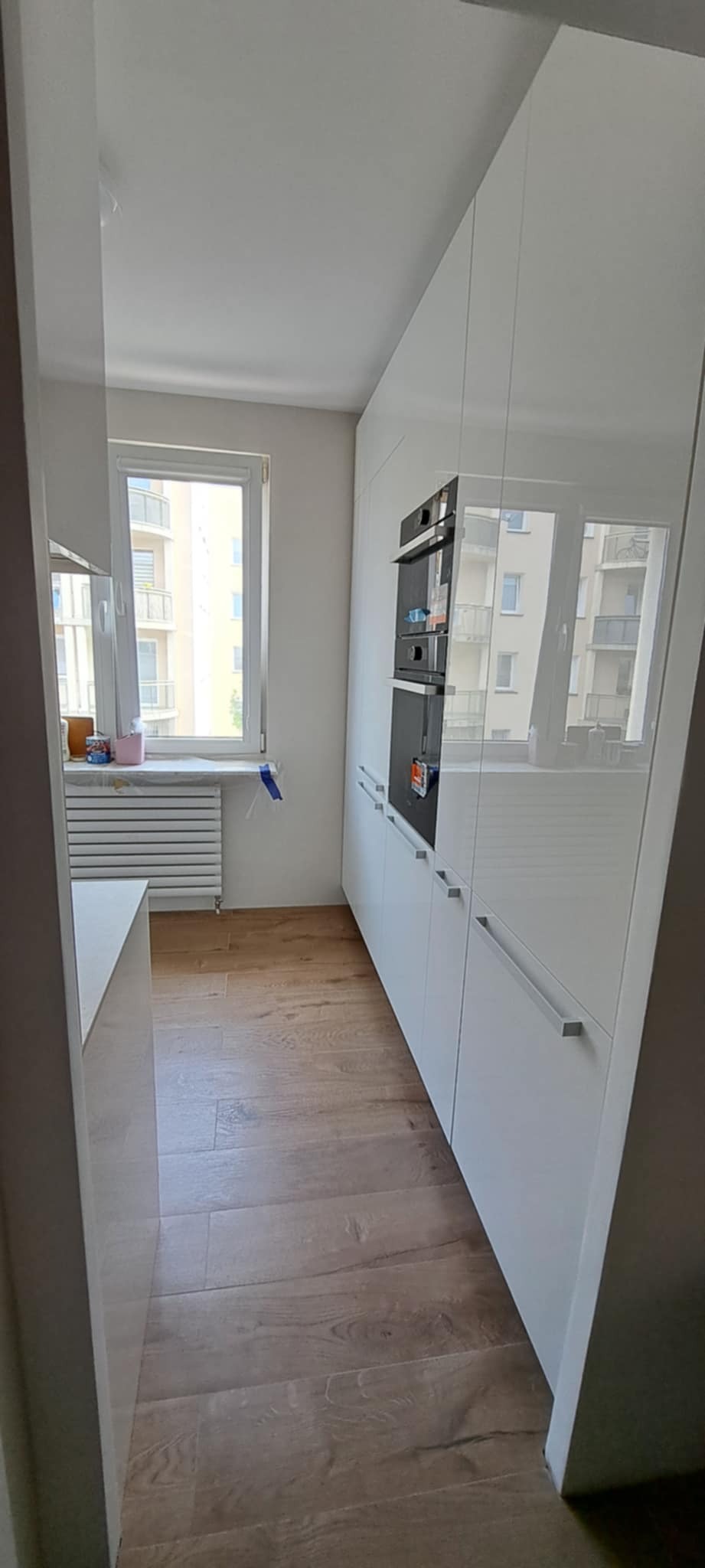 Nowoczesna kuchnia z białymi, lakierowanymi szafkami, drewnianą podłogą i wbudowanym piekarnikiem. Widok na okno z budynkiem w tle. Minimalistyczny design.