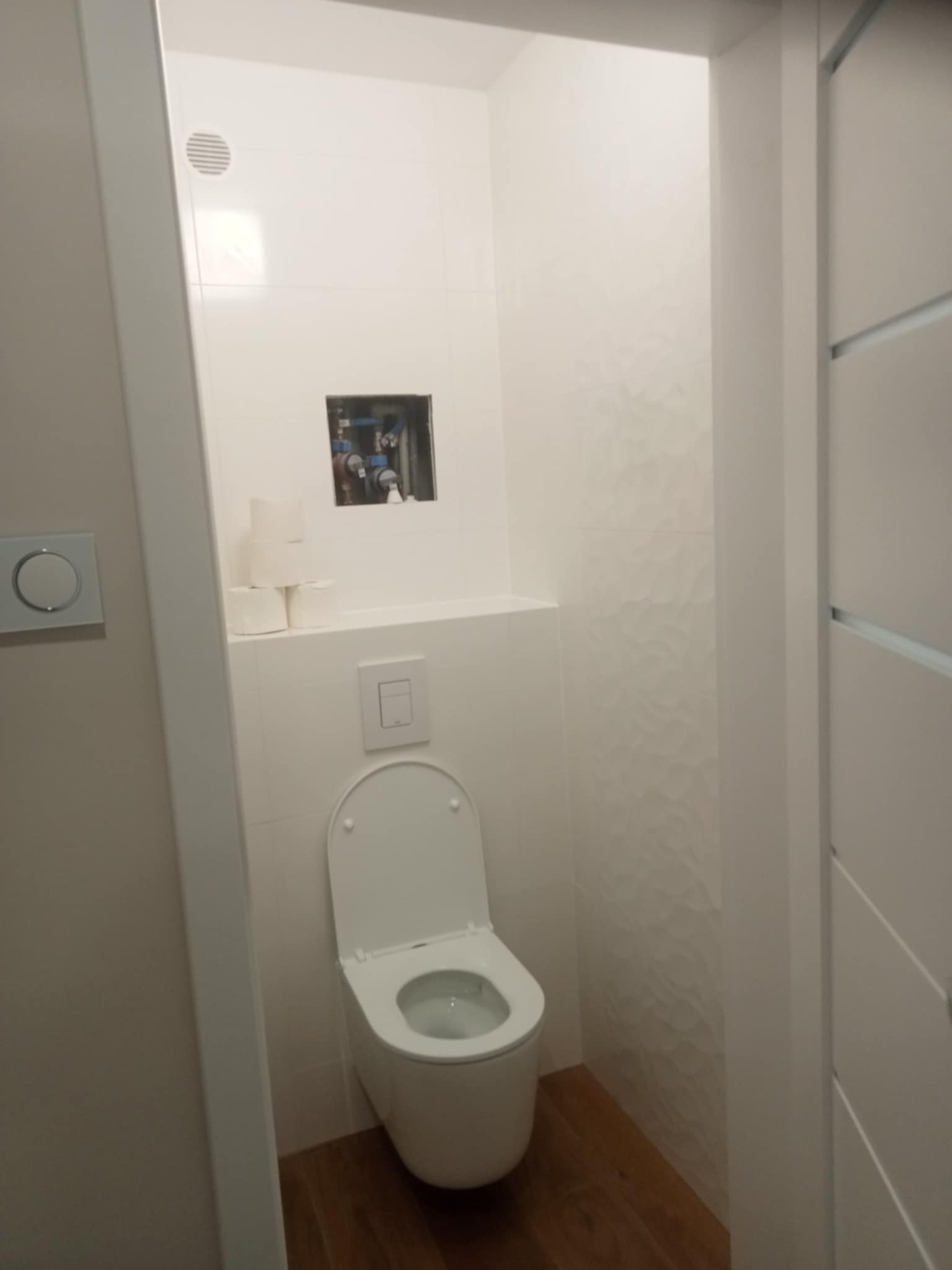 Nowoczesna toaleta w Dmosinie po remoncie. Białe płytki z subtelnym wzorem, podwieszana miska WC i drewnopodobna podłoga. Widoczne instalacje w ścianie.