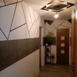 Korytarz z geometrycznymi wzorami w kolorze czarnym na białych ścianach, szary panel ścienny, drewniane drzwi i podłoga, dekoracyjne oświetlenie sufitowe, regał z roślinami w tle.