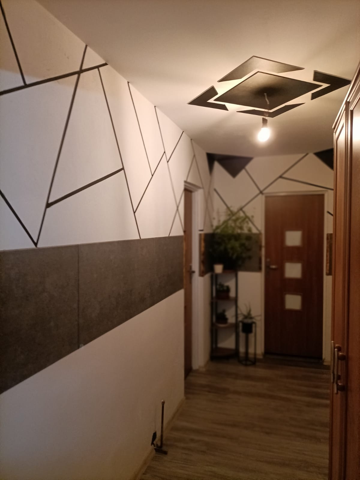 Korytarz z geometrycznymi wzorami w kolorze czarnym na białych ścianach, szary panel ścienny, drewniane drzwi i podłoga, dekoracyjne oświetlenie sufitowe, regał z roślinami w tle.