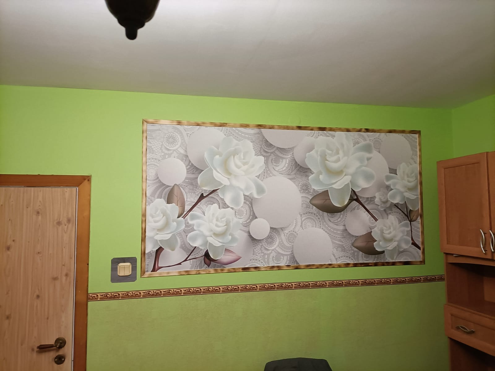 Fragment pokoju z zielonymi ścianami i fototapetą z motywem białych kwiatów w ramie, widoczny fragment drewnianej szafki i drzwi.