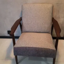 Odrestaurowany fotel w stylu retro z drewnianymi podłokietnikami, tapicerowany tkaniną w odcieniach beżu i szarości, widoczny na tle szarej ściany.