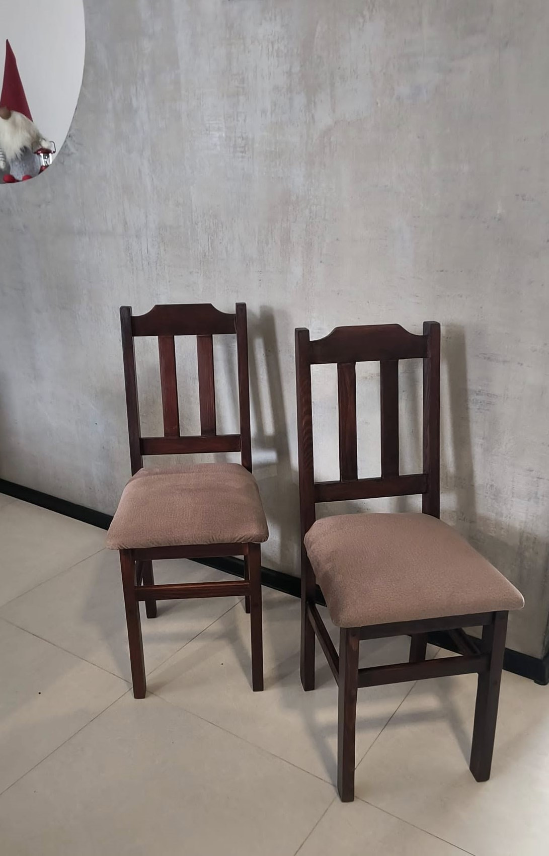 Dwa ciemnobrązowe krzesła z tapicerowanymi siedziskami w odcieniach beżu stoją na tle szarej ściany z widoczną strukturą i fragmentu dekoracji świątecznej.