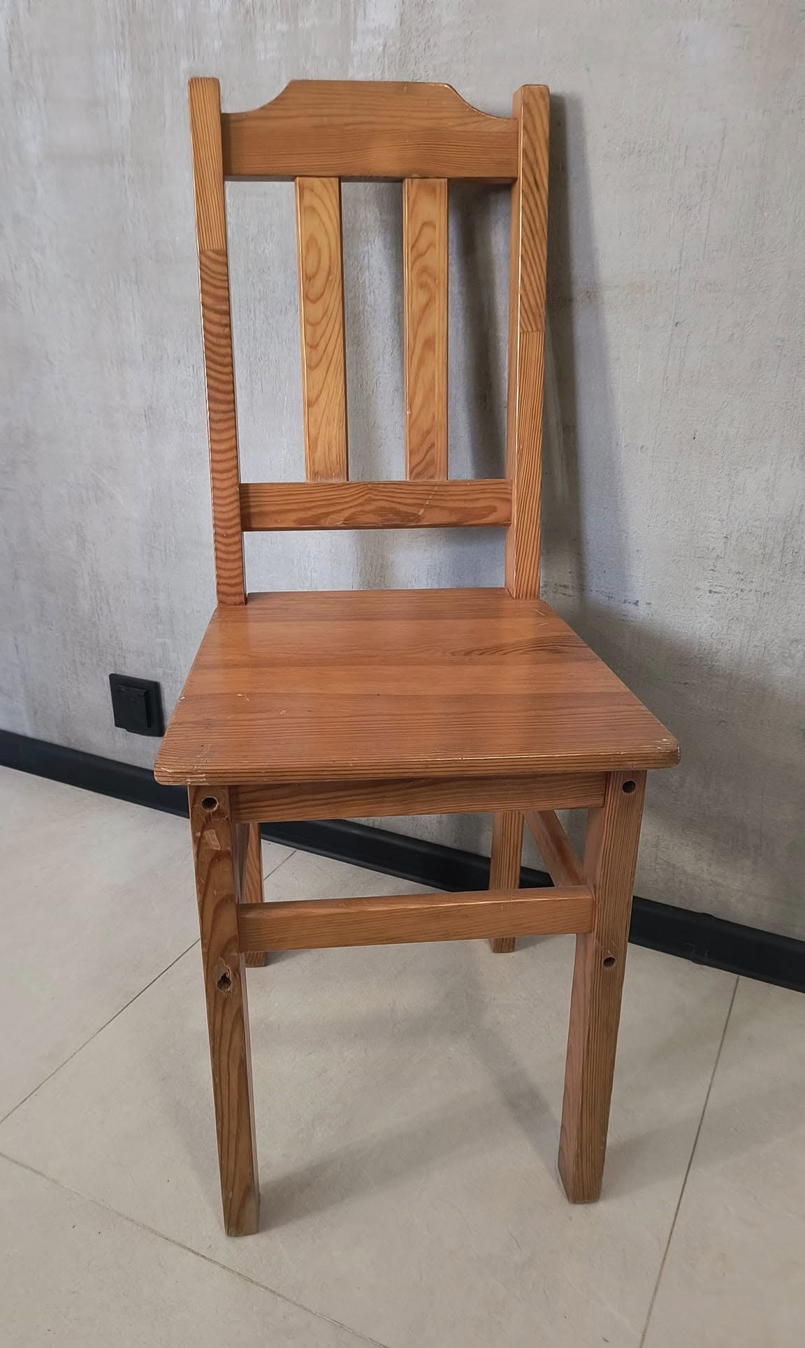 Drewniane krzesło po renowacji, widoczne ślady po wcześniejszych otworach, na tle szarej ściany z czarną listwą przypodłogową.