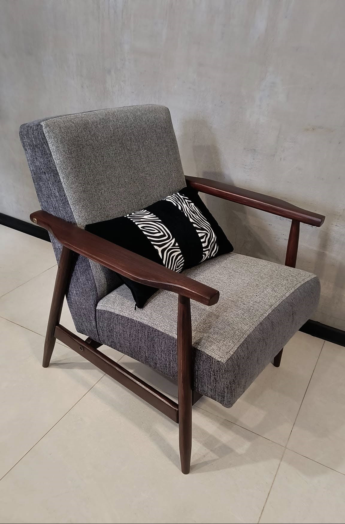 Odrestaurowany fotel w stylu retro z ciemnego drewna i szarą tapicerką, z poduszką w czarno-białe pasy, na tle betonowej ściany.