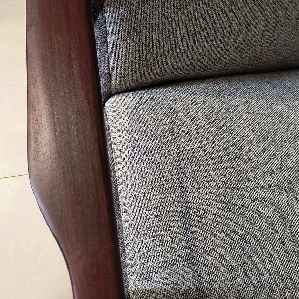 Fragment odnowionego fotela z ciemnobrązowym, lakierowanym drewnianym podłokietnikiem i szarą, grubą tkaniną obiciową o wyraźnym splocie.