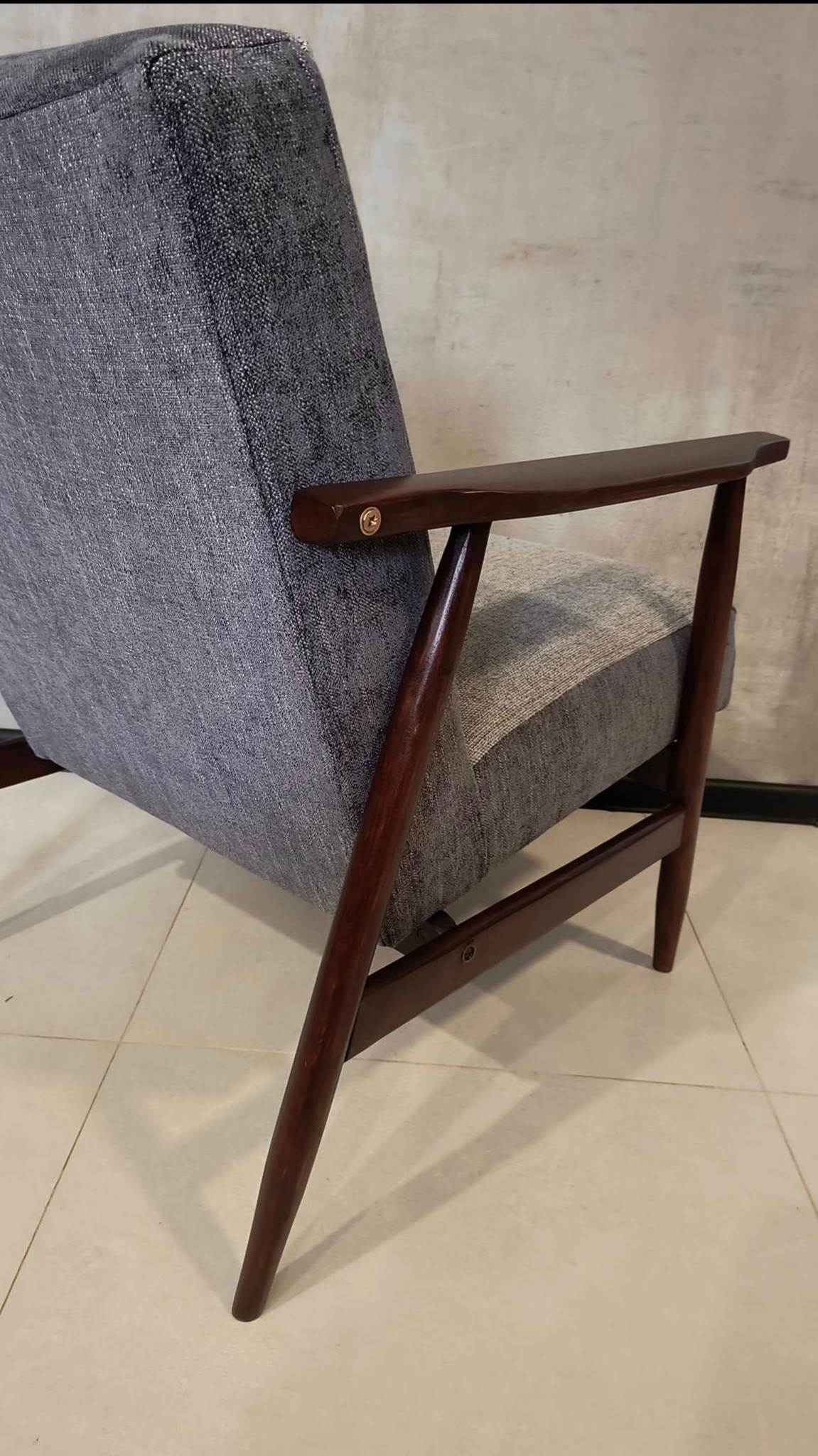 Odrestaurowany fotel w stylu vintage z szarą tapicerką i ciemnobrązowymi drewnianymi nóżkami, widok z boku, z widocznym mocowaniem śrubowym na podłokietniku.