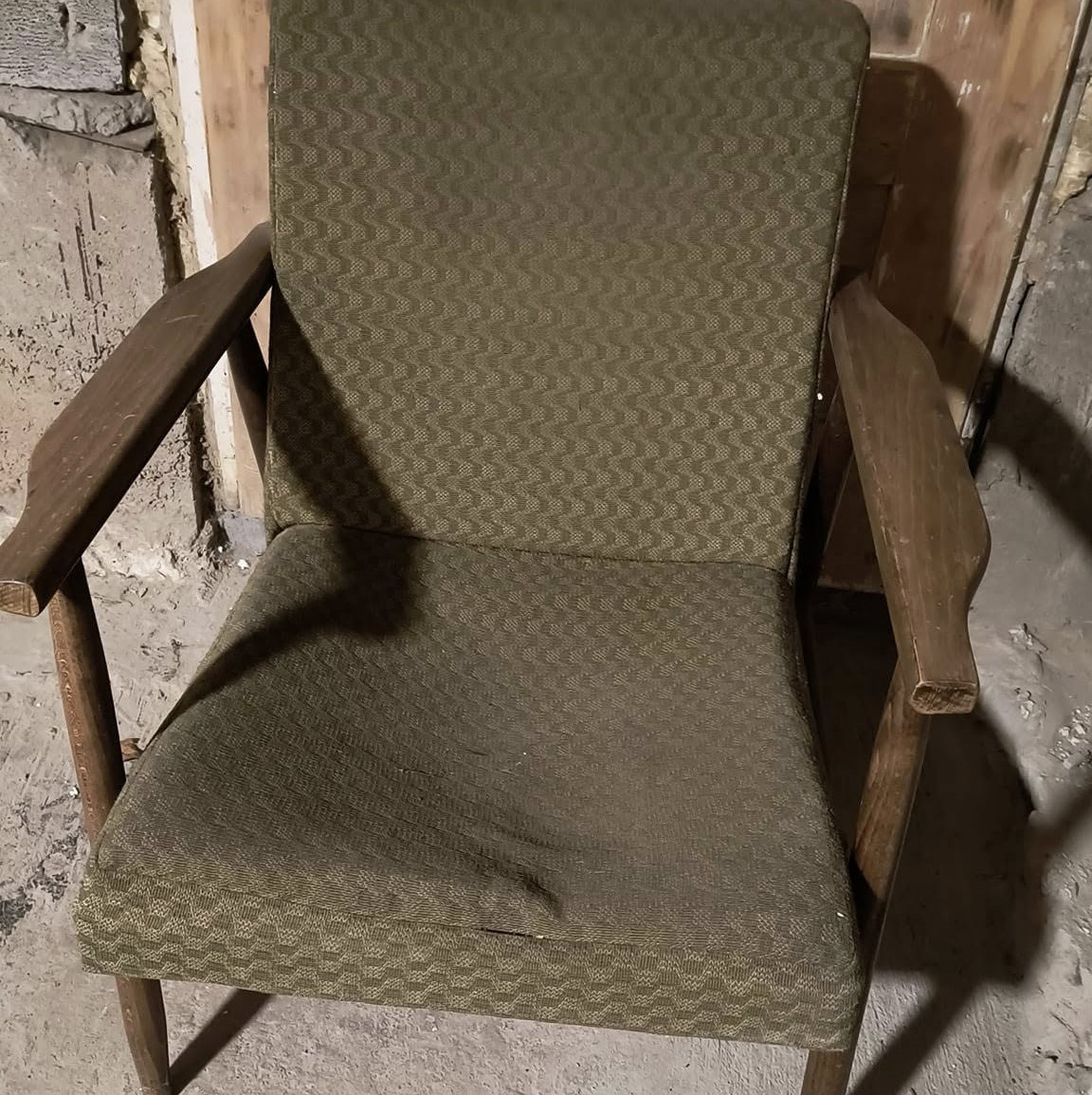 Stare krzesło z drewnianymi podłokietnikami i zieloną tapicerką w geometryczny wzór, widoczne uszkodzenia na siedzisku i zabrudzenia, tło stanowią surowe ściany i podłoga.