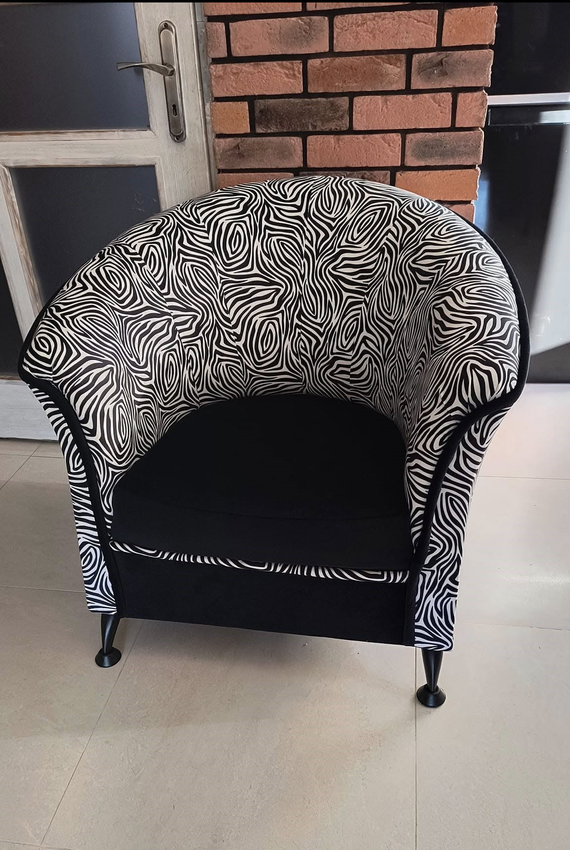 Fotel tapicerowany w czarno-biały wzór zebry, z czarnym siedziskiem, stojący na jasnych płytkach, z fragmentem ściany z cegły w tle.