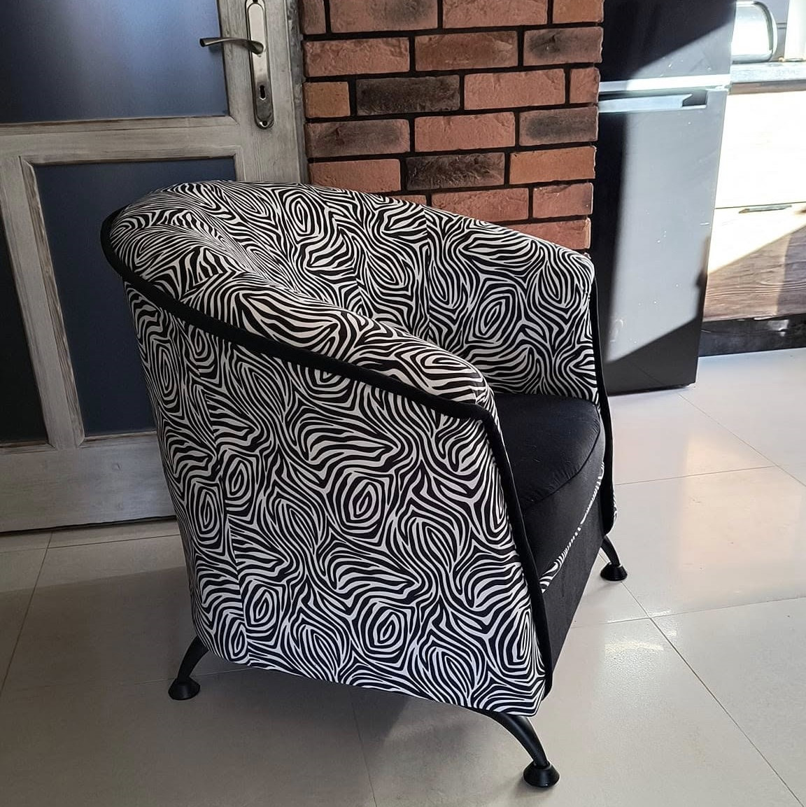 Kubełkowy fotel po renowacji, obity tkaniną w czarno-biały wzór zebry, z czarnymi nóżkami i lamówką, stojący na białej podłodze w pomieszczeniu z ceglaną ścianą.