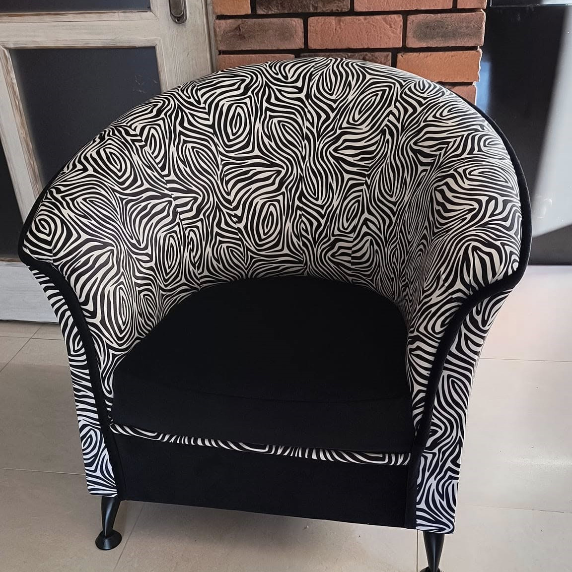Fotel klubowy po renowacji, obity tkaniną w czarno-białe wzory przypominające zebrę, z czarnym siedziskiem i nóżkami, na tle ceglanej ściany i białych drzwi.