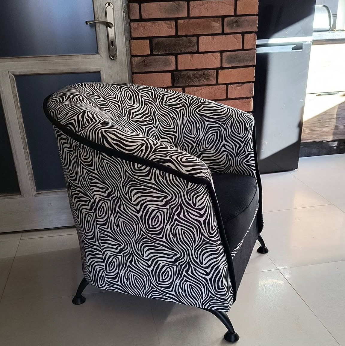 Tapicerowane krzesło z czarno-białym wzorem zebry, czarne obicie siedziska, czarne nóżki, widoczne na tle ceglanej ściany i szarej szafki.